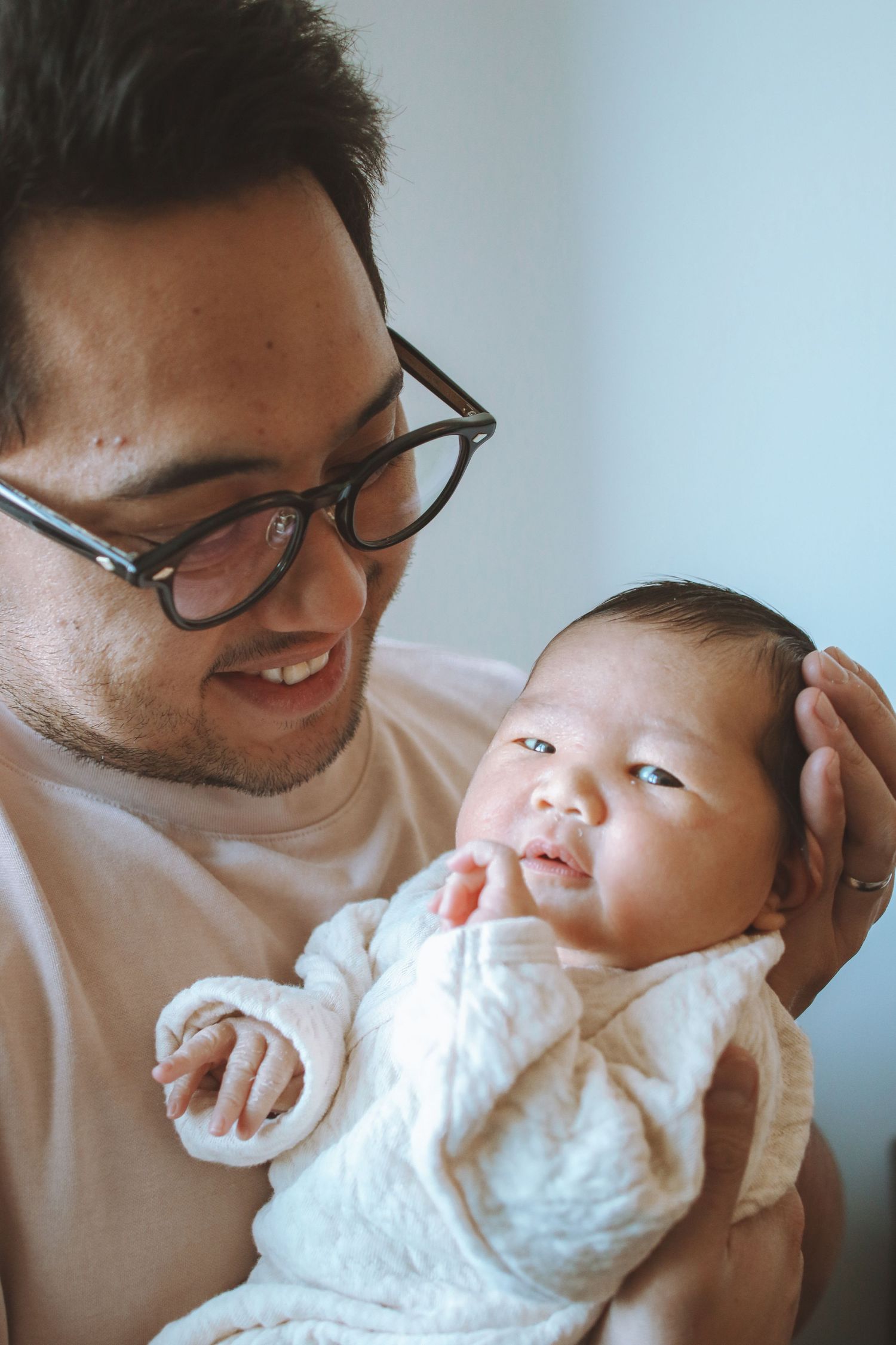 Proud father with glasses smiles while holding alert newborn baby wrapped in white.