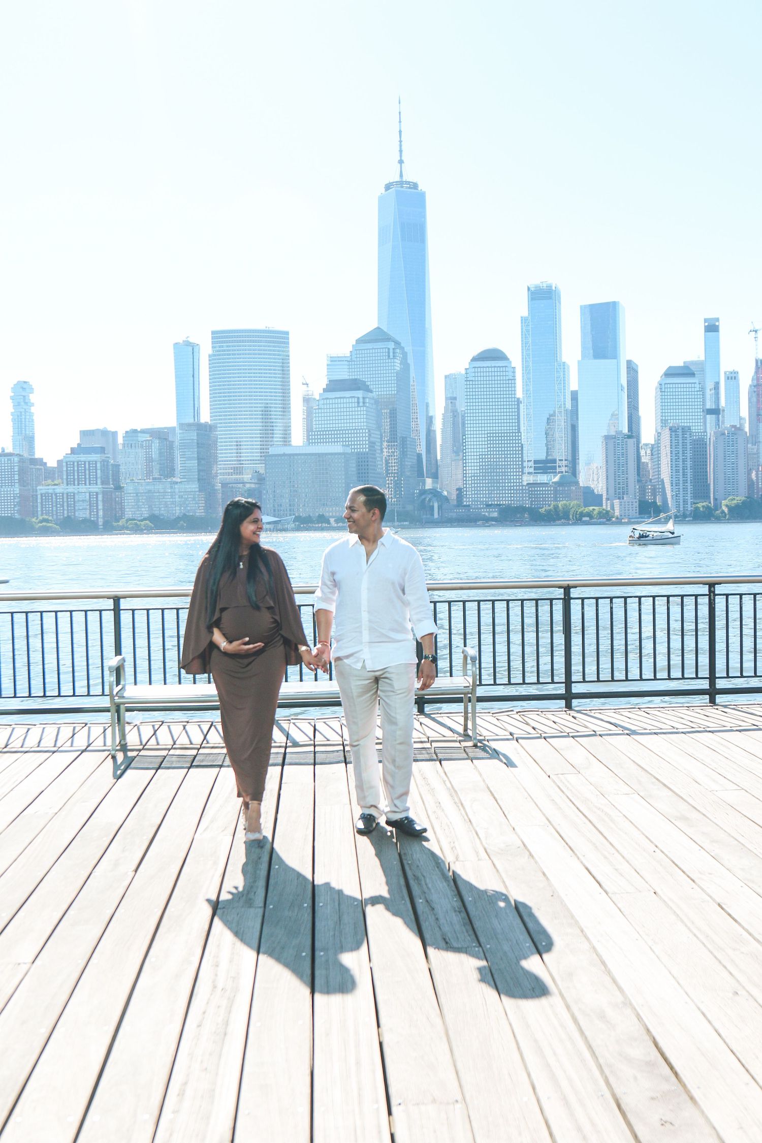 Pregnant couple holding hands on a waterfront boardwalk with the New York City skyline in the background.