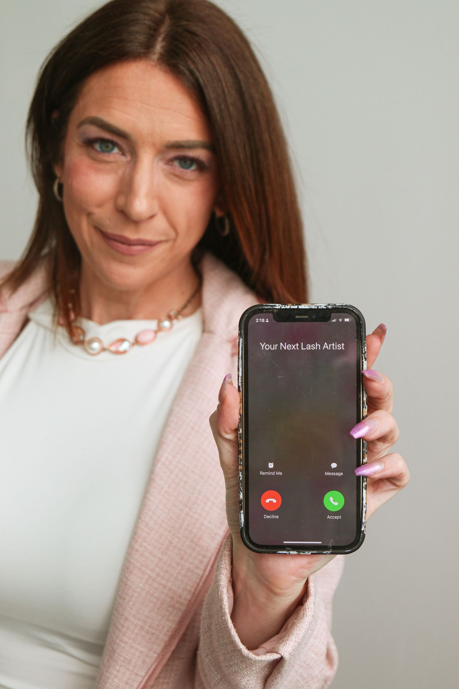 Woman in pink blazer holding phone showing incoming call from Your Next Lash Artist.