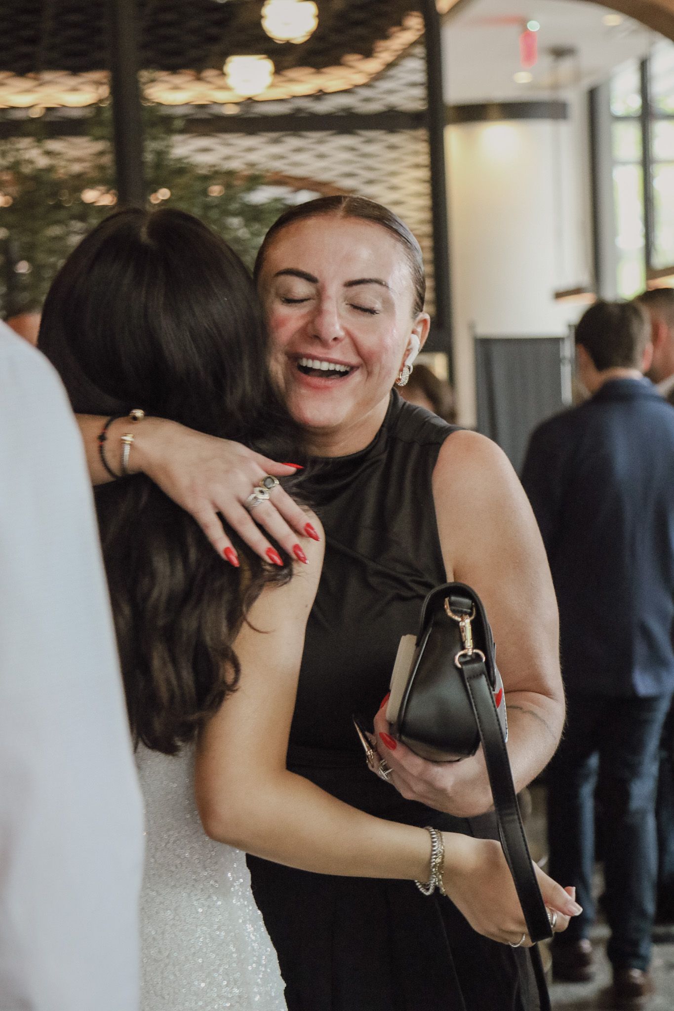A smiling woman in a black sleeveless dress with a handbag embraces another woman in a white dress at an indoor gathering.