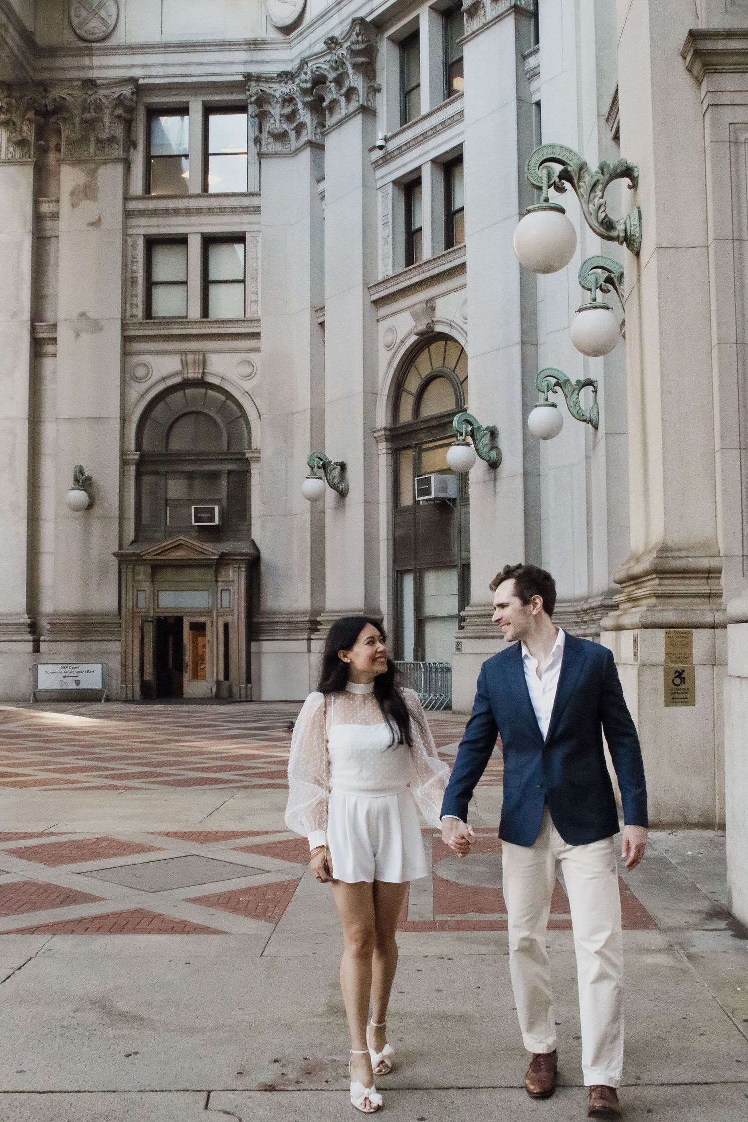 Newlywed couple walks hand in hand past ornate NYC building facade, bride in white romper, groom in navy blazer.