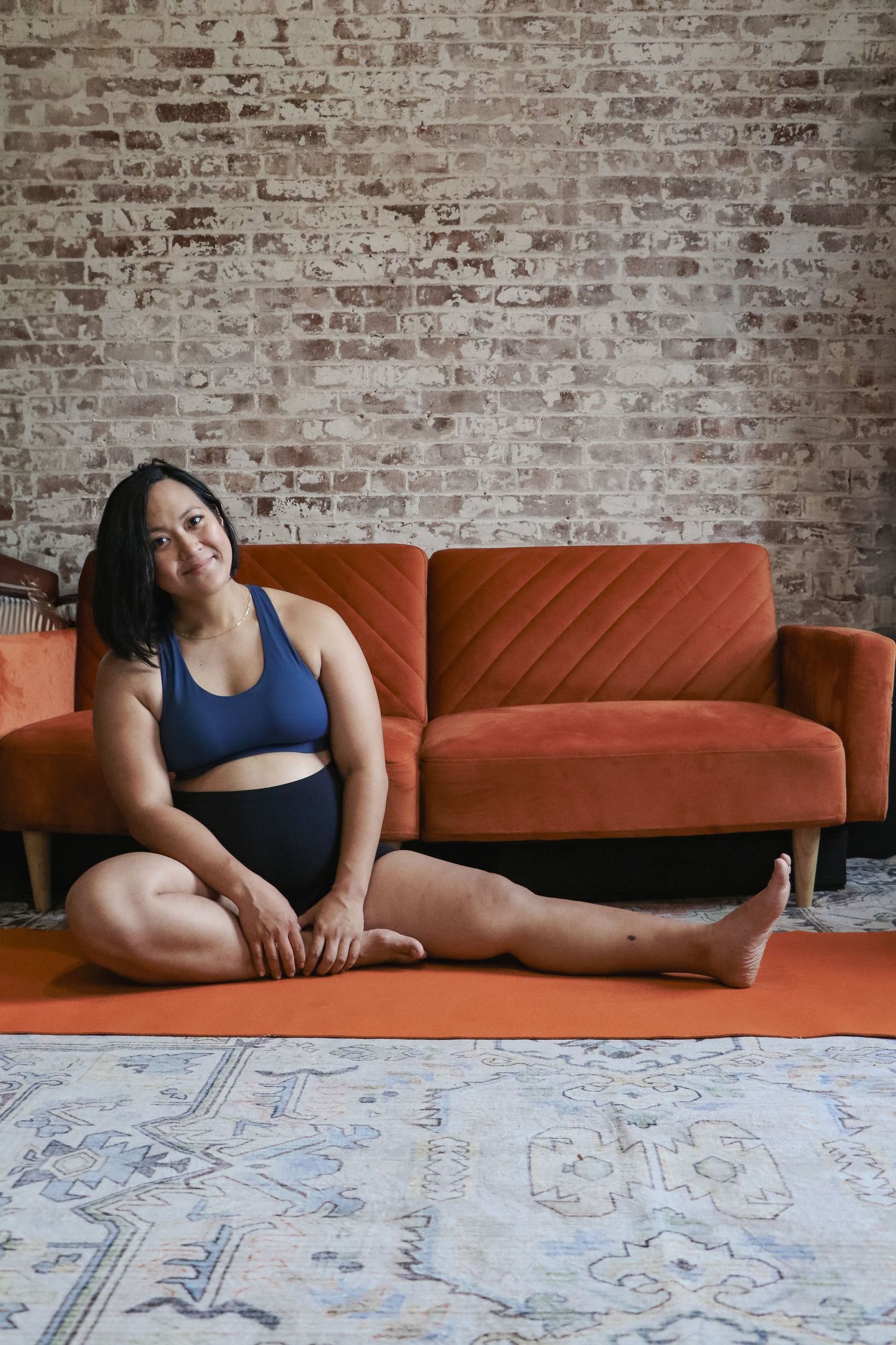Smiling woman in blue sports bra and black shorts sitting on orange yoga mat near red velvet sofa.