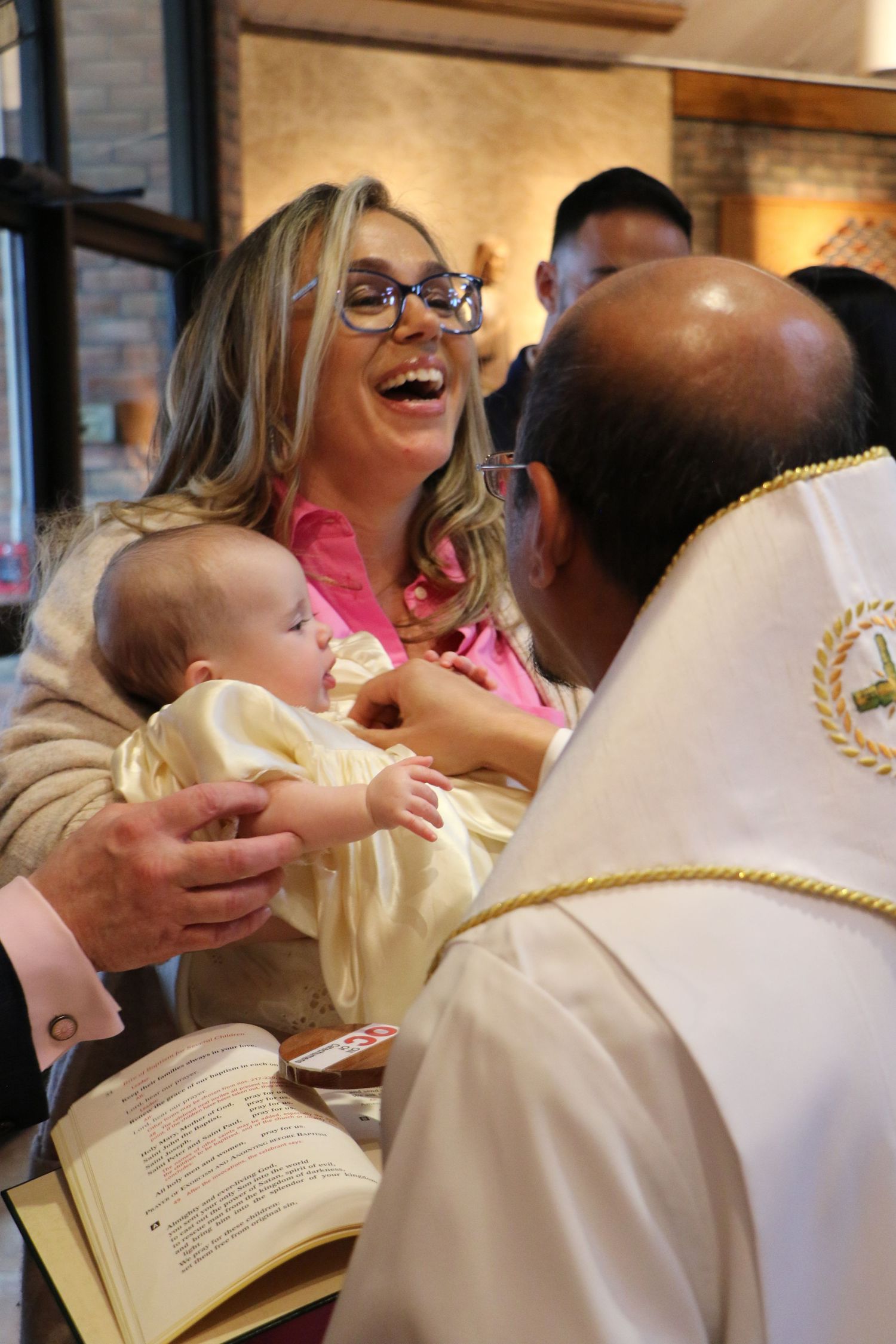 Laughing woman with glasses holding baby at a religious baptism ceremony with priest.