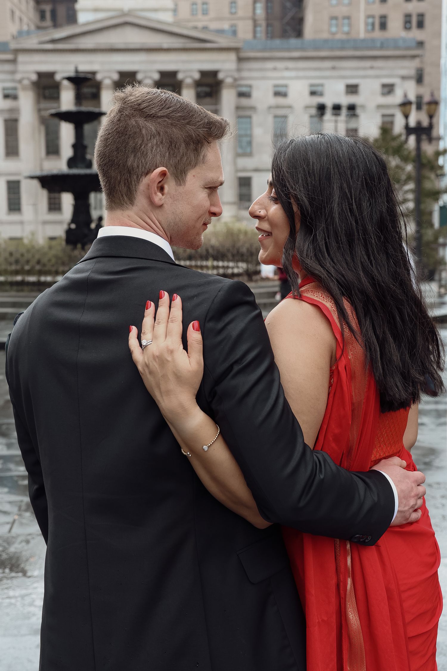 Couple embracing in front of a stately columned building and fountain, the woman in a red dress with red nails and the man in a black suit.