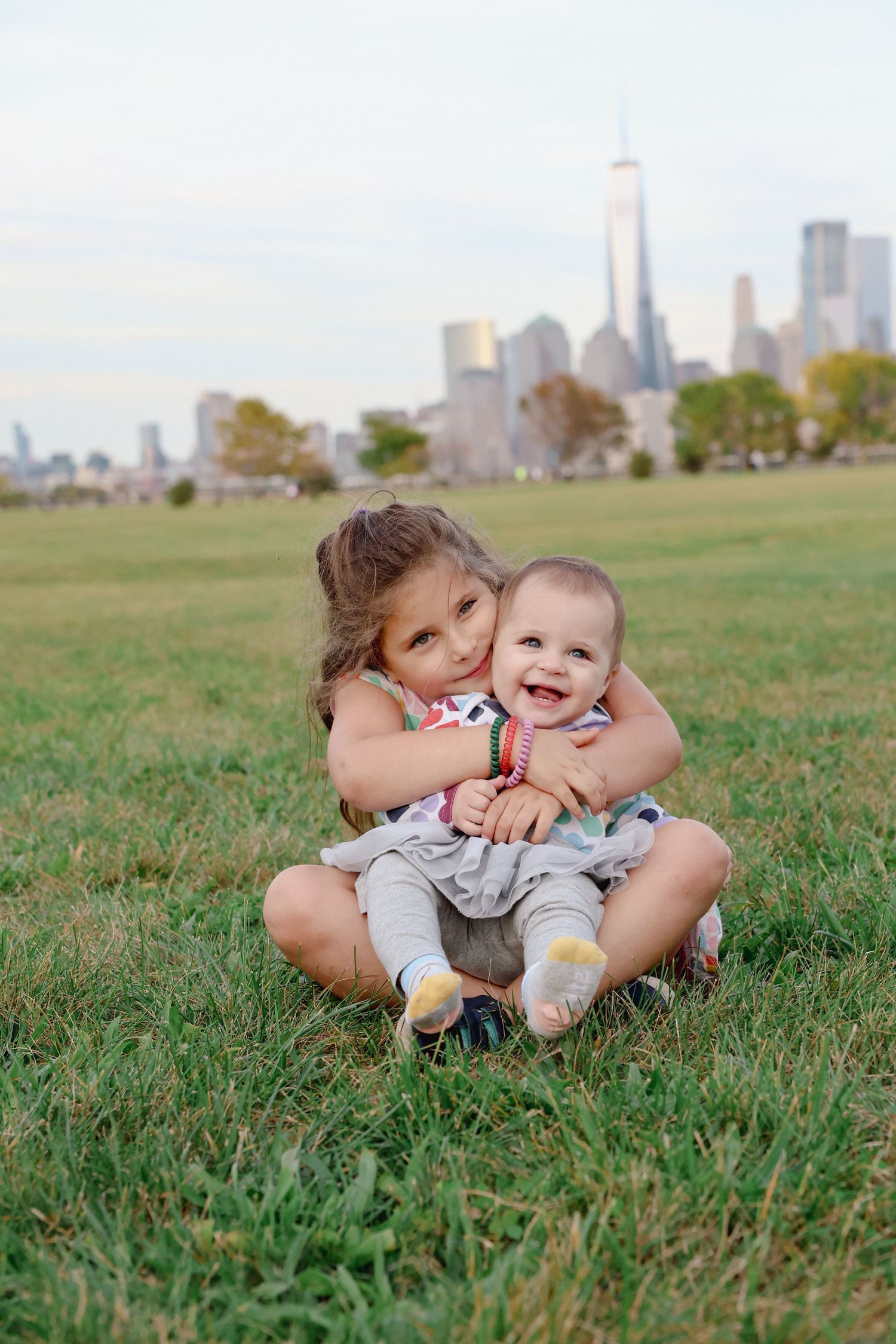 Young girl hugging a smiling baby while sitting on a grassy lawn with a city skyline in the background.