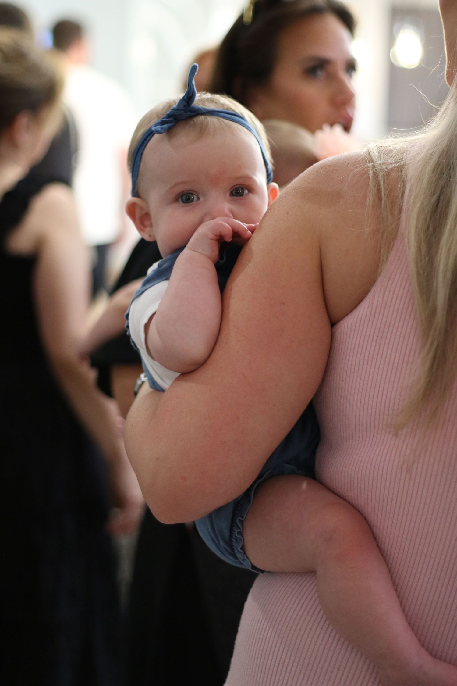 A baby with a blue headband being held by a woman at an indoor event.