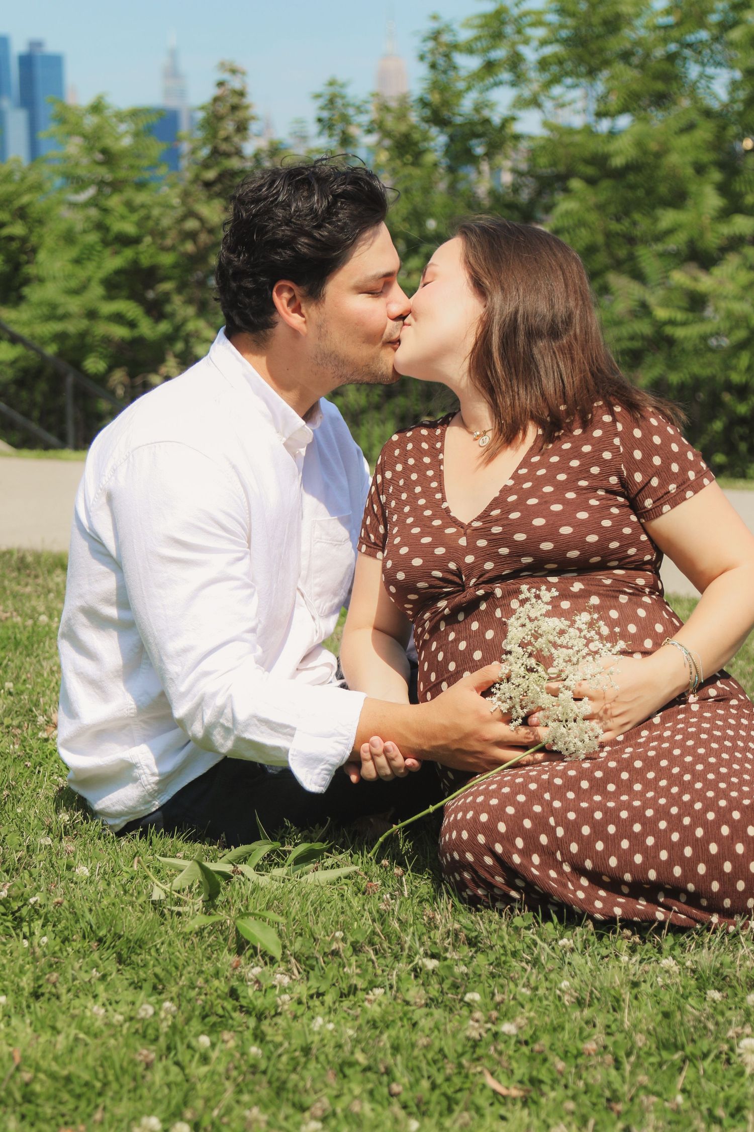 Pregnant couple kissing on grass in park, woman in brown polka dot dress holding white flowers on sunny day.