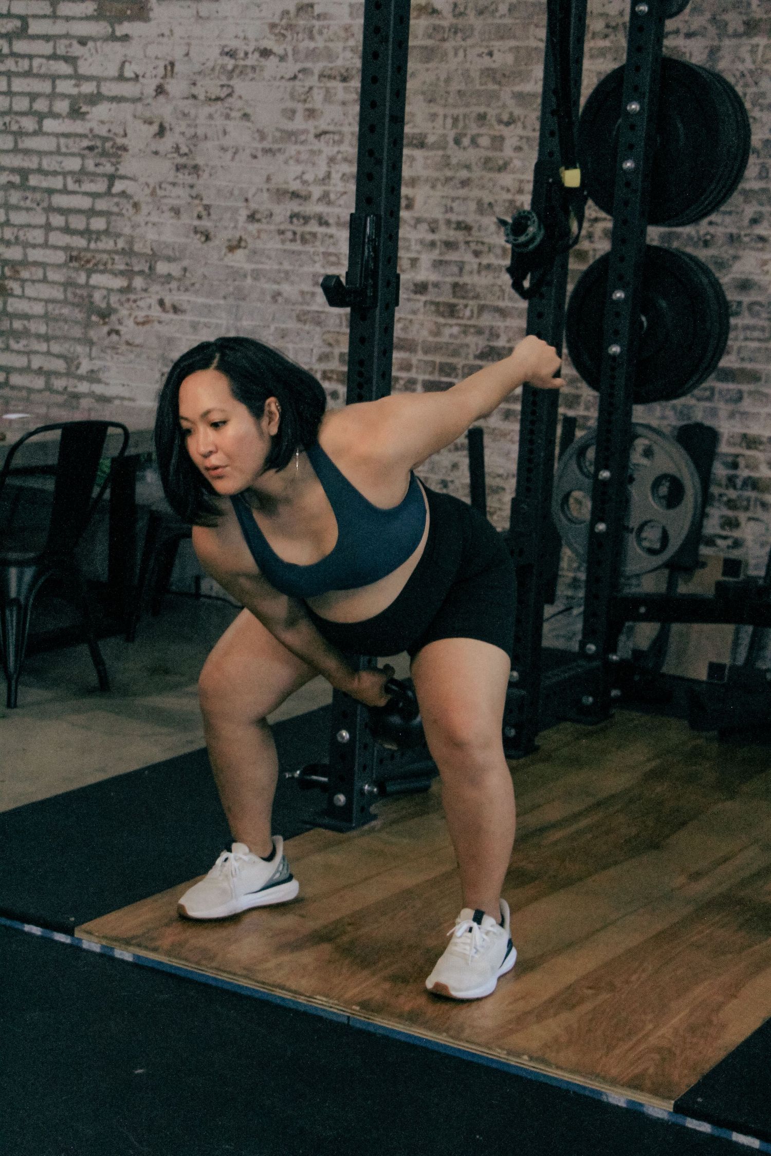 Woman performing kettlebell deadlift in gym with brick wall and squat rack background.