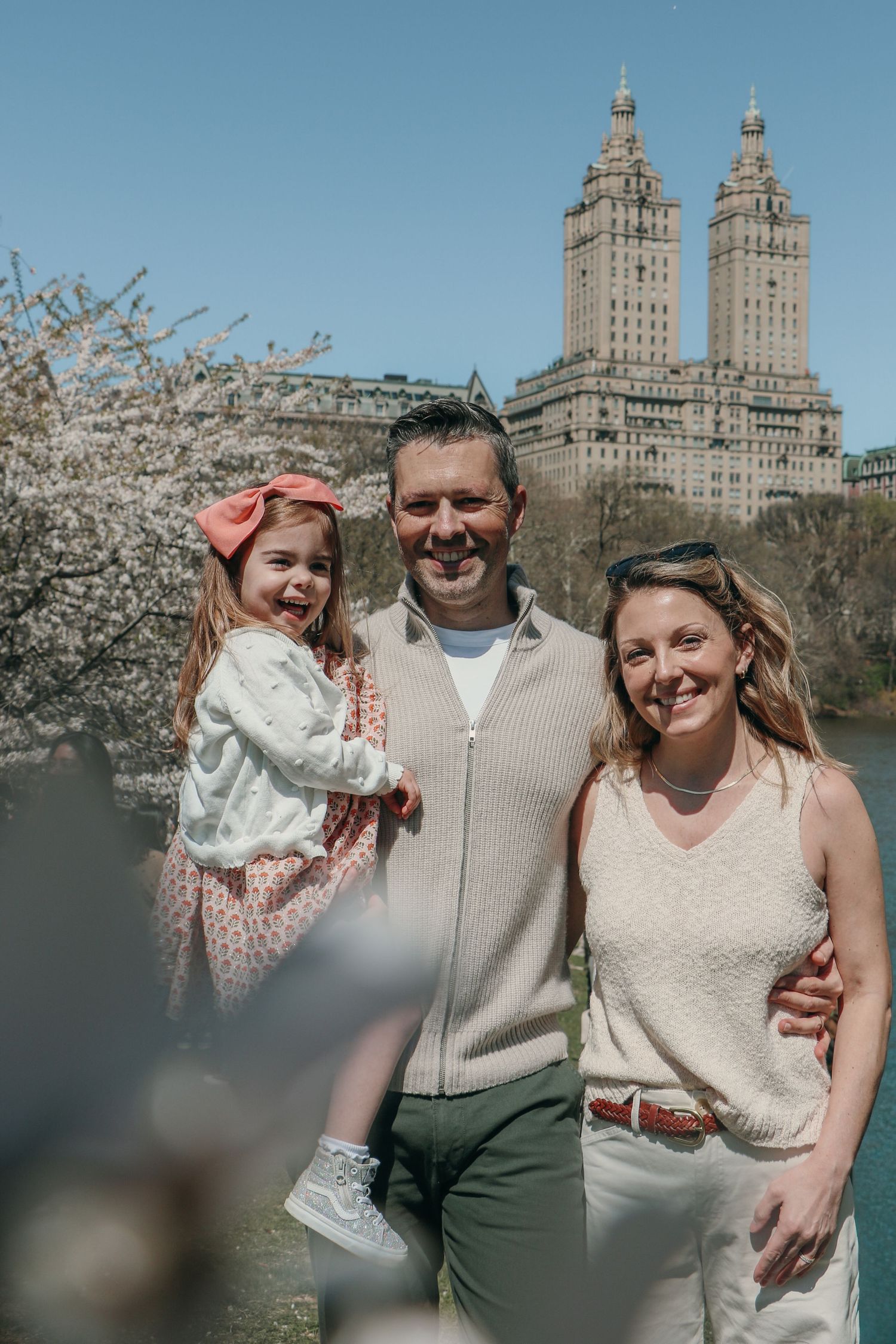 Happy family with toddler girl poses near cherry blossoms with Central Park buildings backdrop.