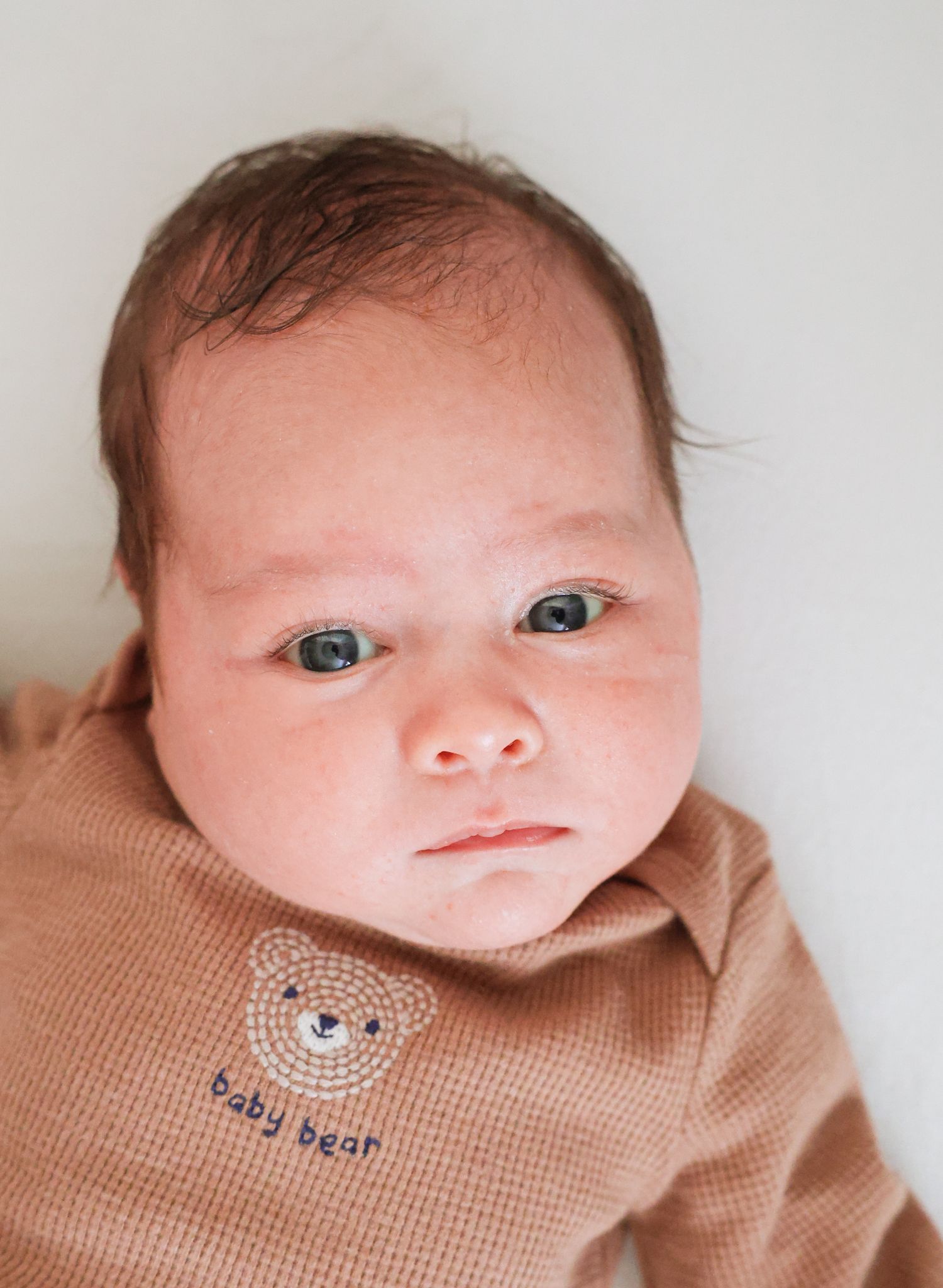 Newborn baby with dark hair and blue-gray eyes wearing a tan waffle-knit top with an embroidered 