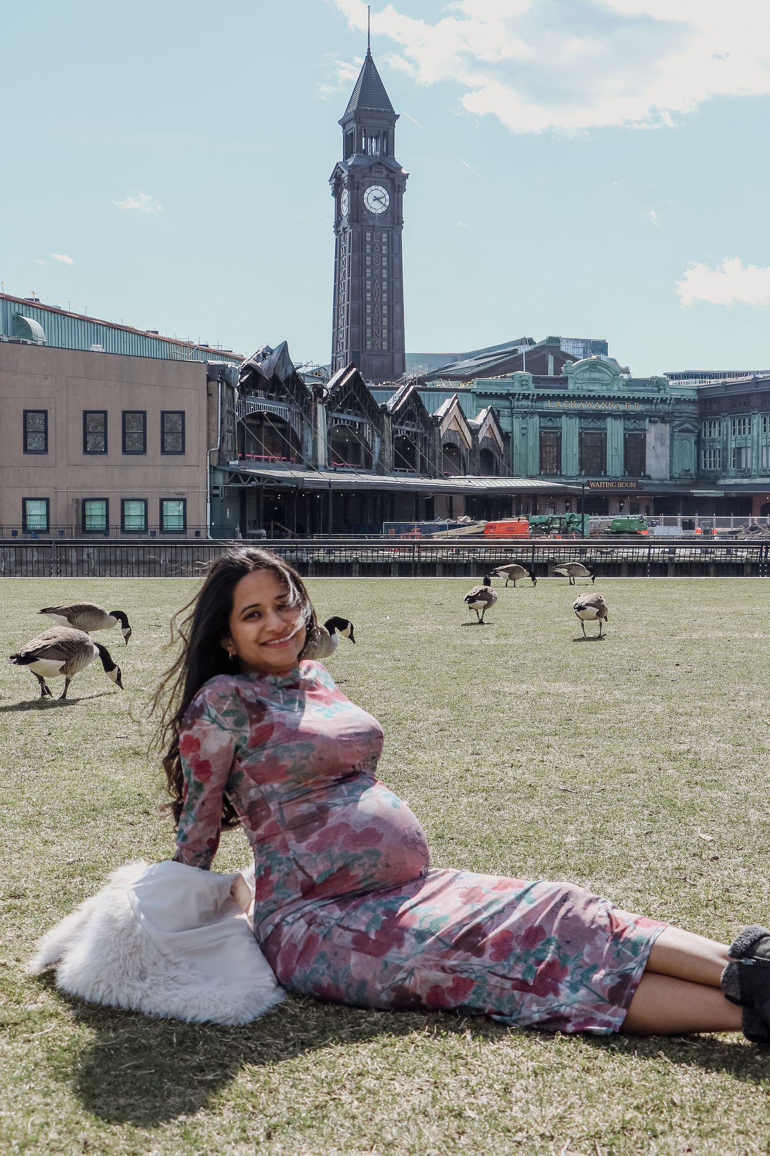 Smiling pregnant woman in floral dress sits on grass near historic clock tower on sunny day.
