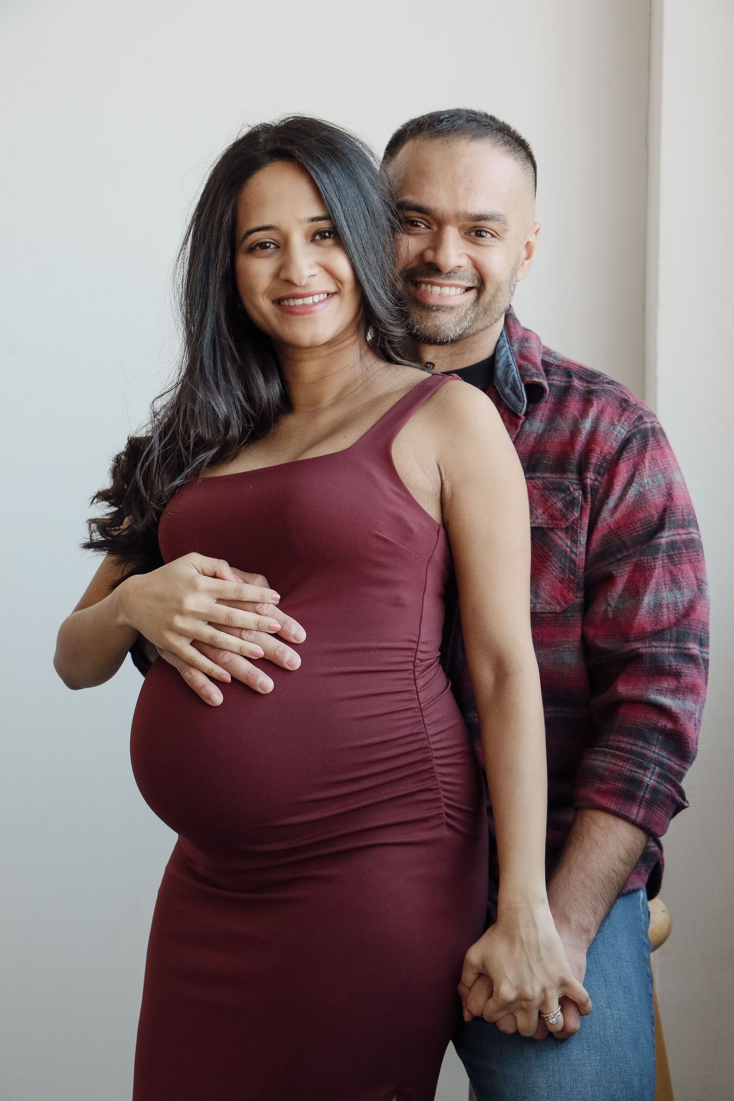 Pregnant woman in burgundy dress with partner behind her, both smiling at camera indoors.