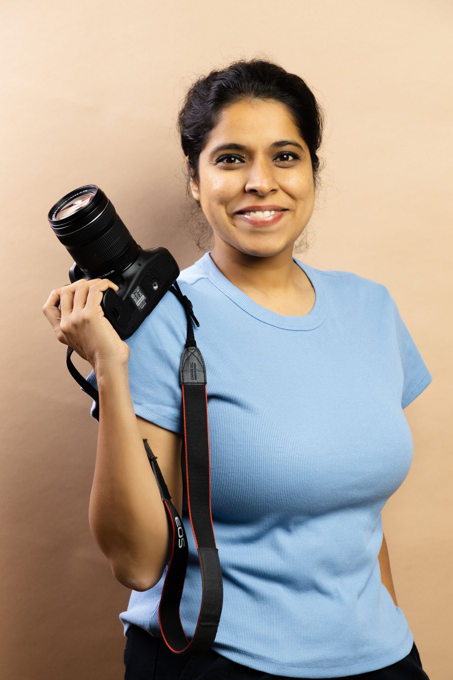 Smiling Indian woman in blue t-shirt holding DSLR camera with strap against beige background.