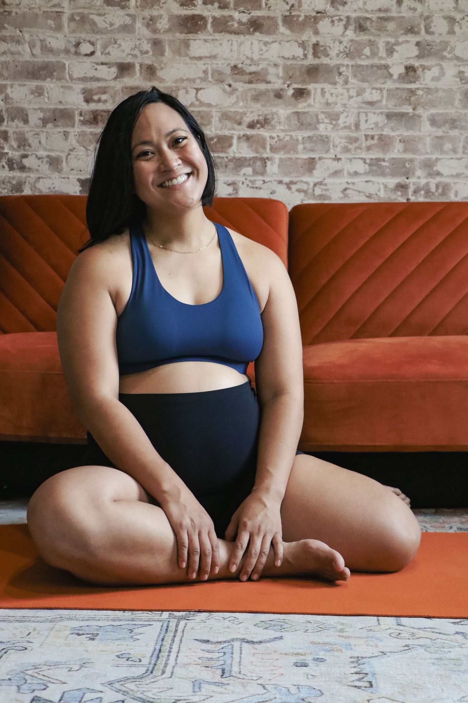 Happy woman in blue workout set sitting cross-legged on an orange yoga mat in front of a red sofa.