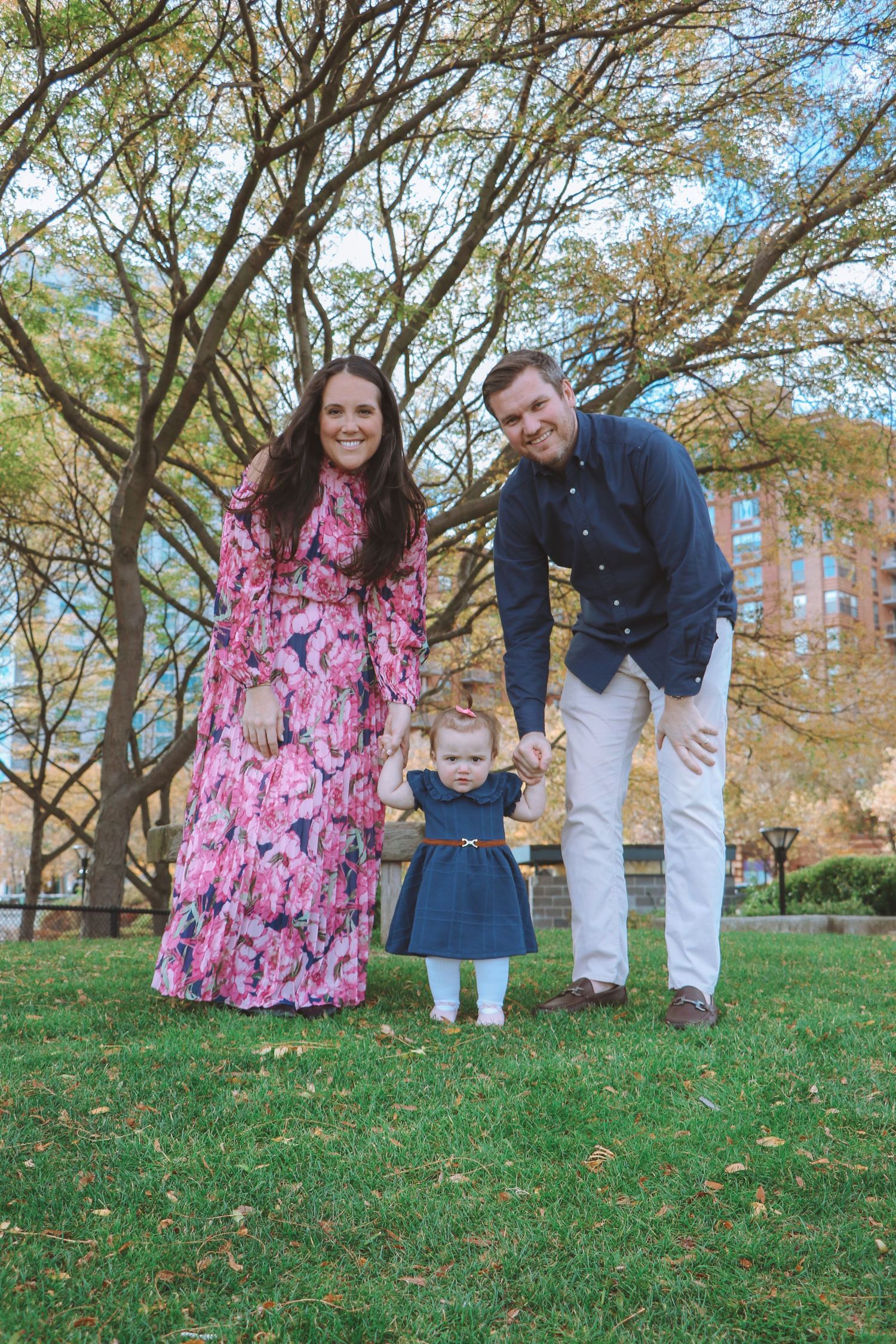 A happy family of three poses outdoors, parents holding toddler girl in navy dress on green grass.