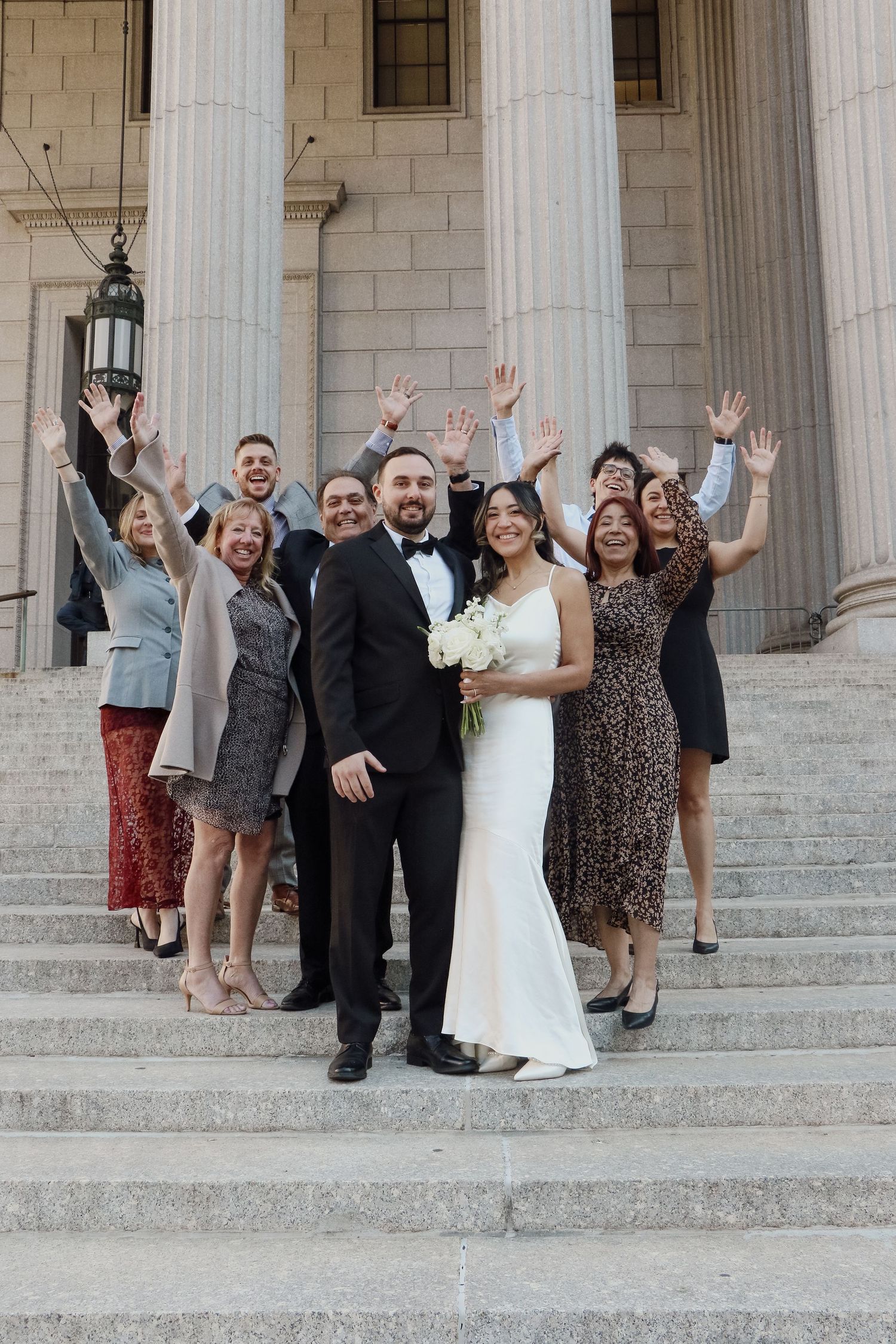 Wedding party celebrates on courthouse steps, raising hands joyfully behind bride and groom in NYC.
