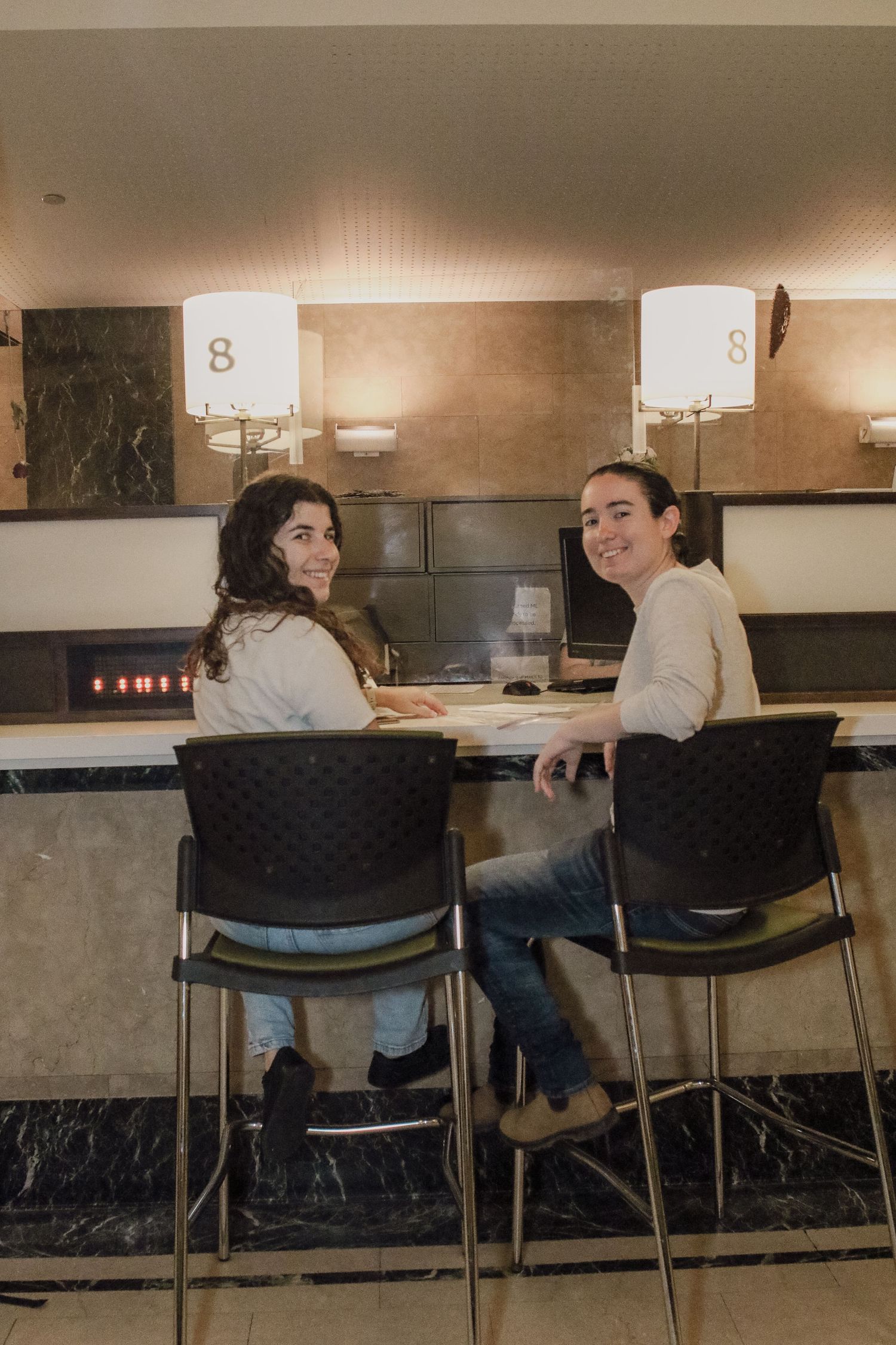 Two women sitting at a bar counter smiling at camera in a warmly lit hotel lobby interior.