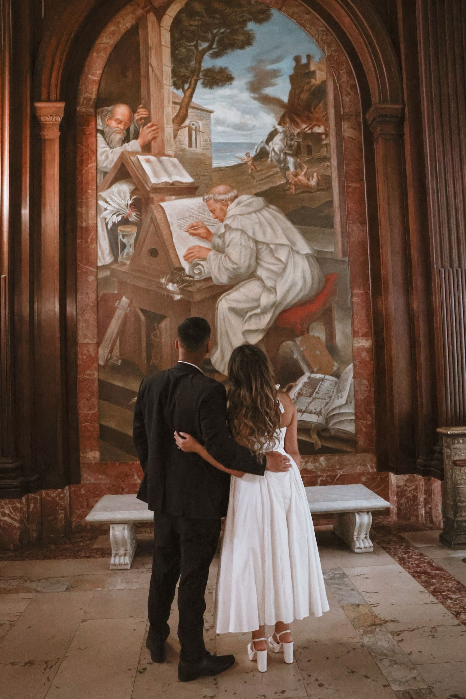Couple in formal attire embracing as they admire a large religious mural of robed monks writing in a historic chapel.