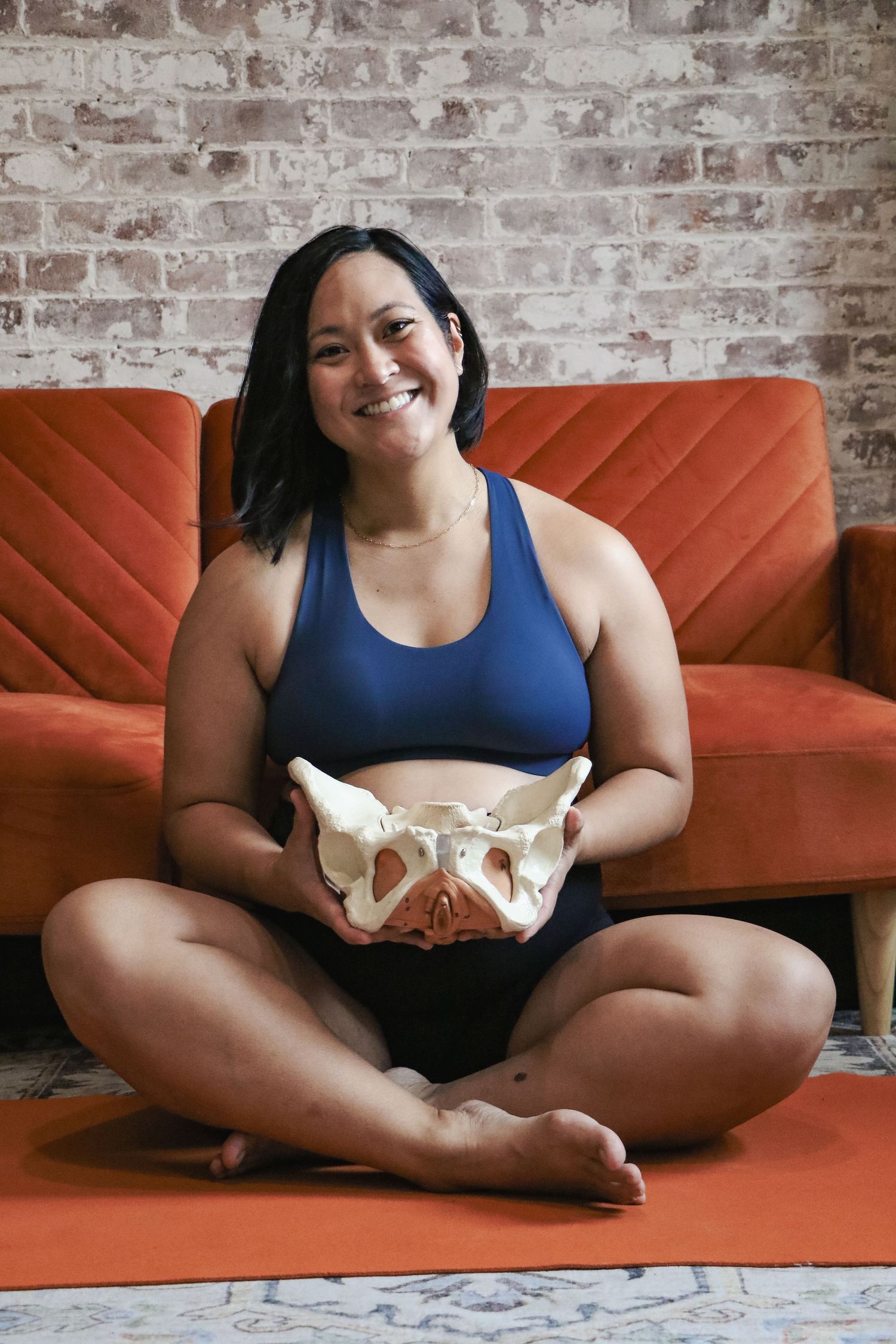 Smiling woman in blue sports bra sitting cross-legged on yoga mat holding a human pelvis bone model.