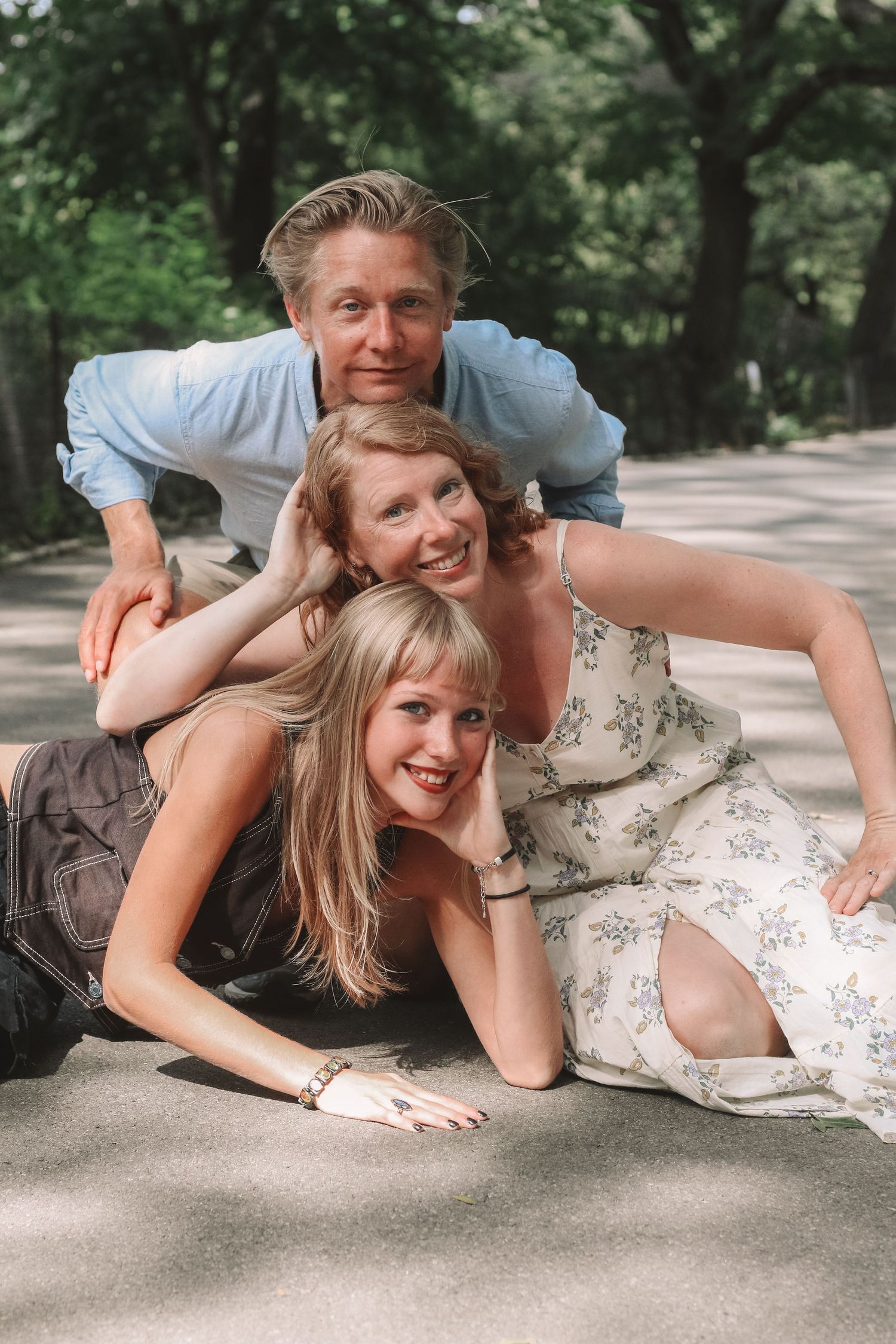 Playful family of three posing stacked on ground in sunny park.