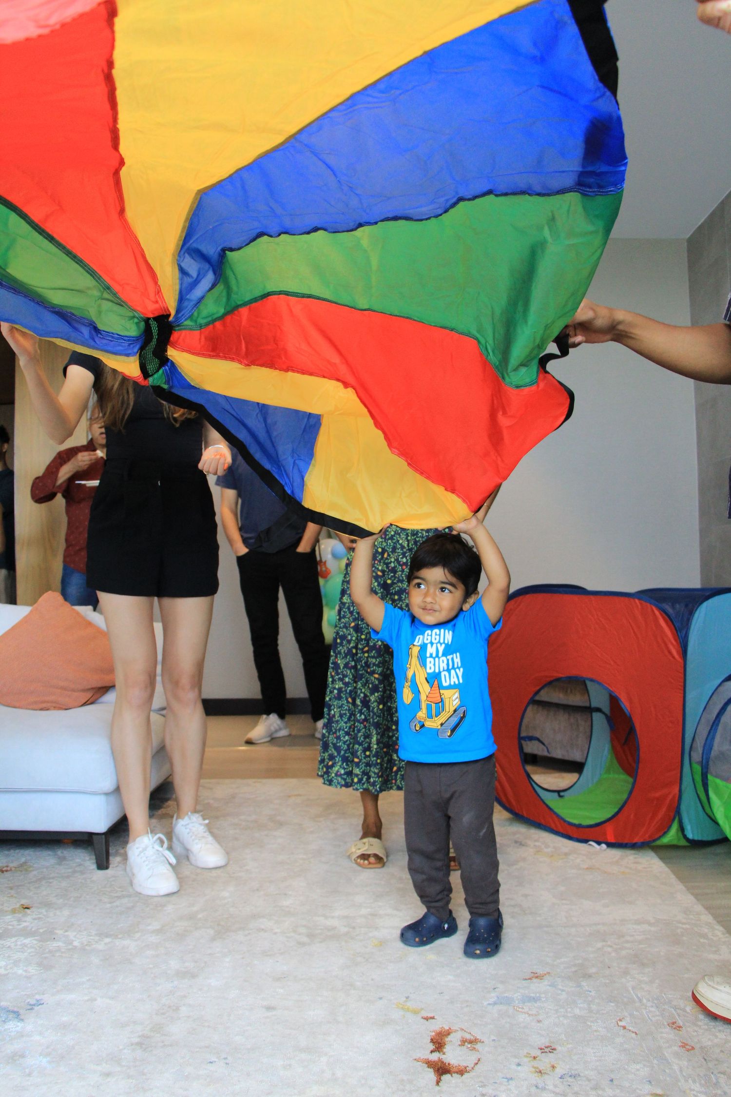 Birthday boy in blue shirt stands under a colorful parachute during indoor birthday party games.