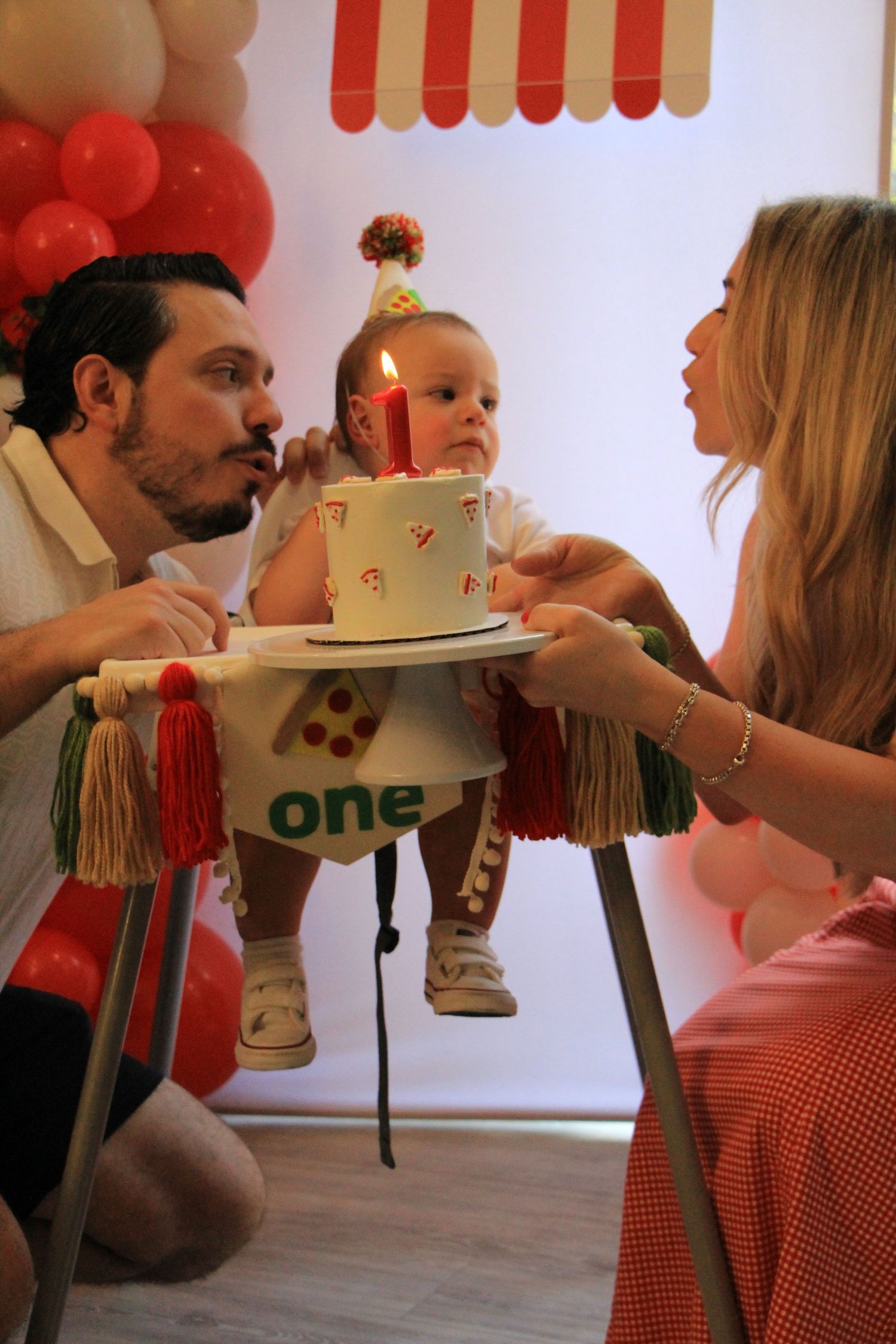 Parents present a first birthday cake with a candle to their baby sitting in a decorated high chair.