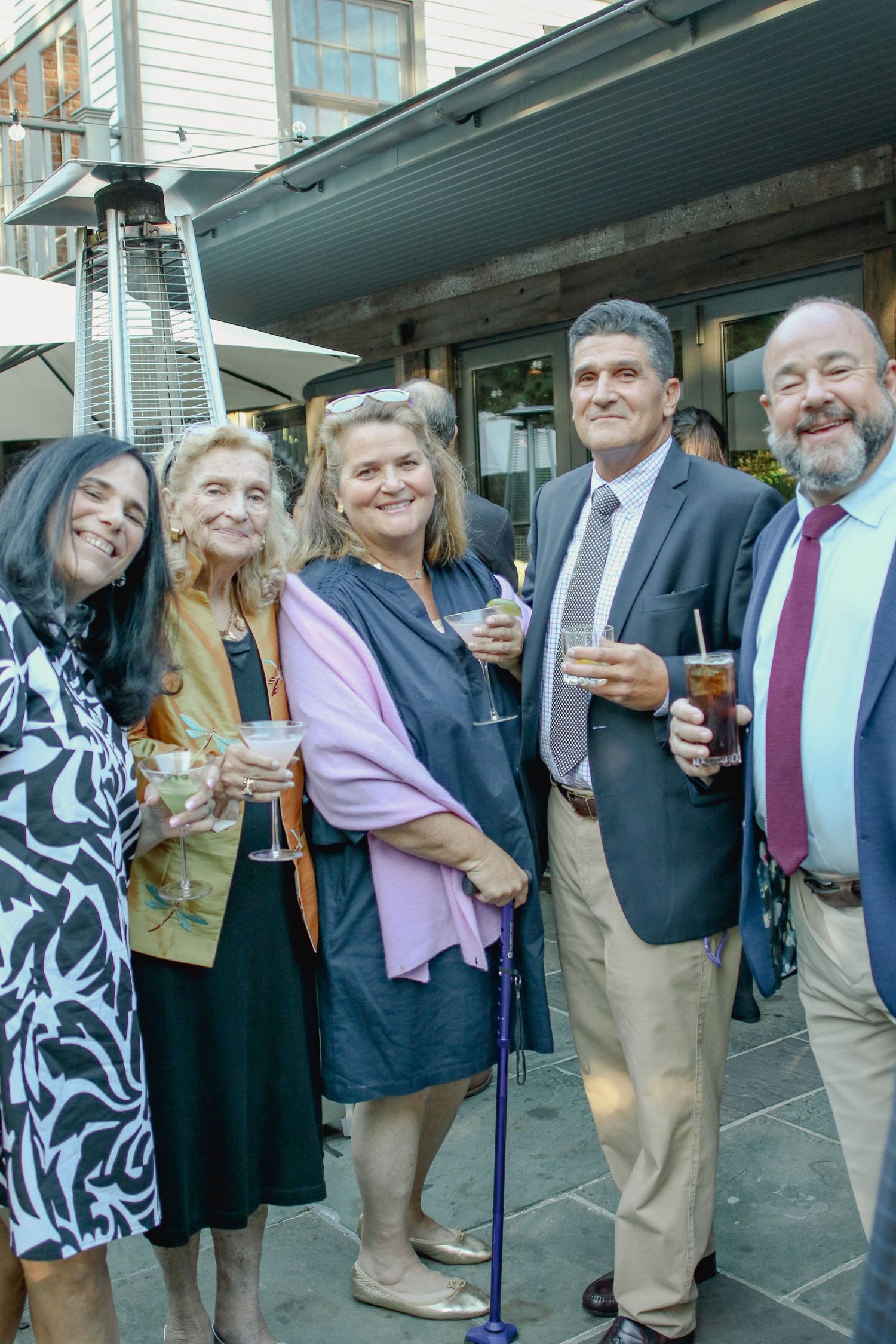 Group of five adults smiling and holding drinks at outdoor evening event.
