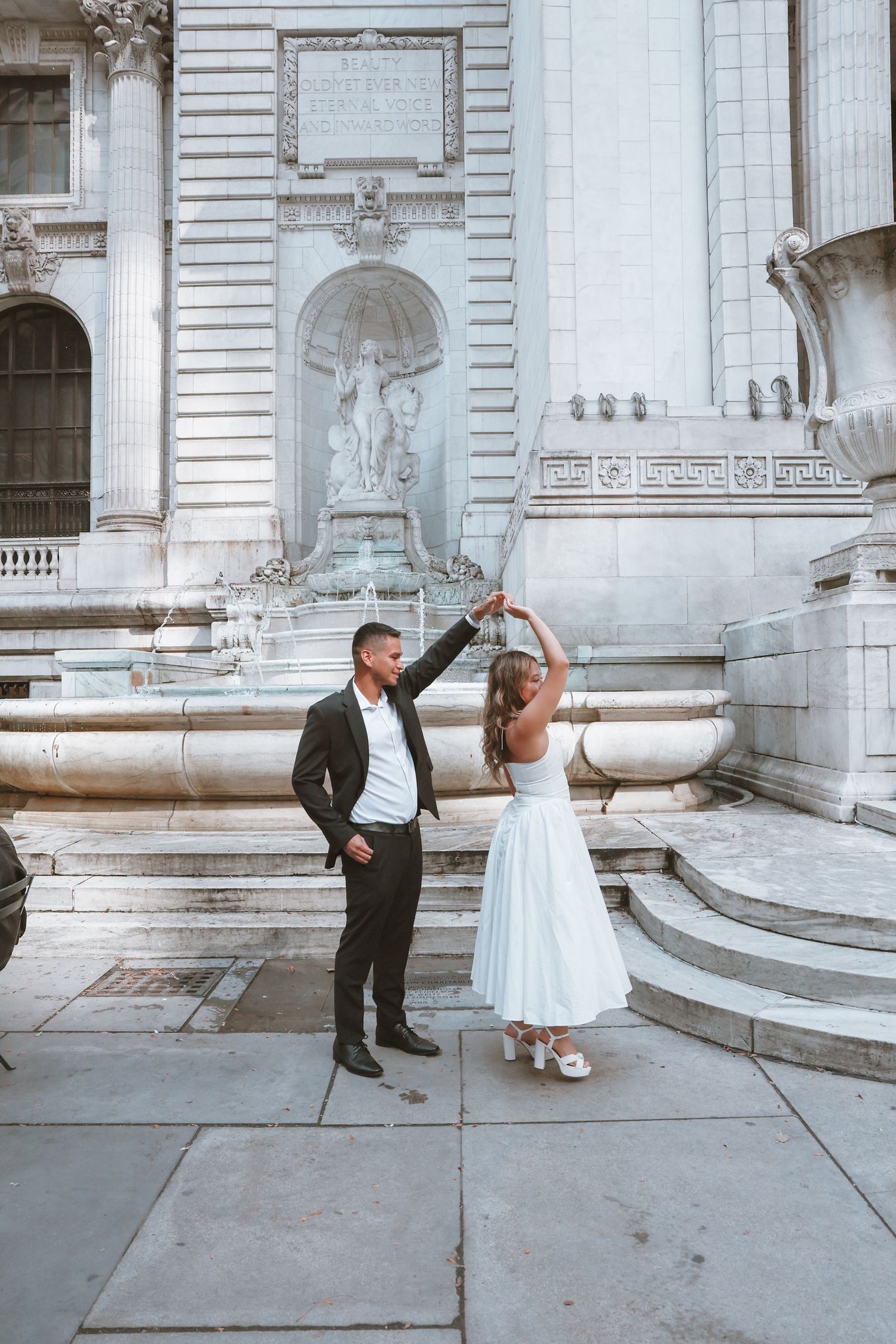 Couple in formal wedding attire dance and twirl on stone steps in front of an ornate marble fountain and neoclassical library building.