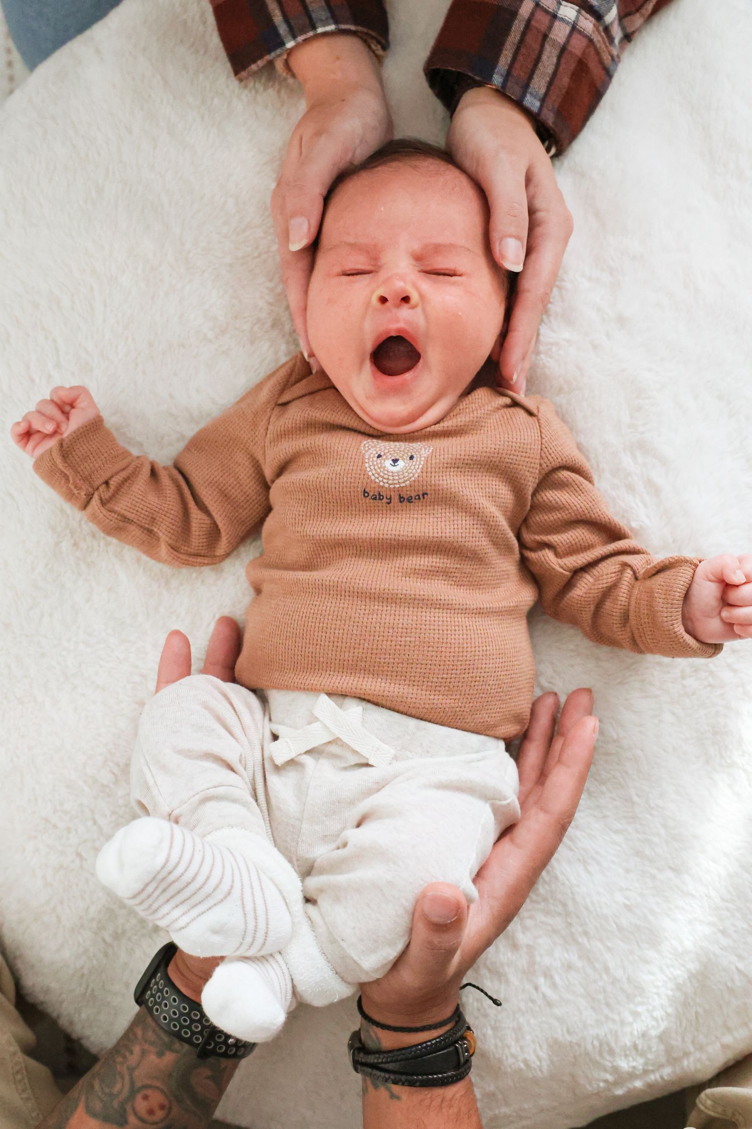 Yawning newborn baby in a tan bear onesie and white pants lies on a cream blanket while adult hands gently cradle the head and feet.