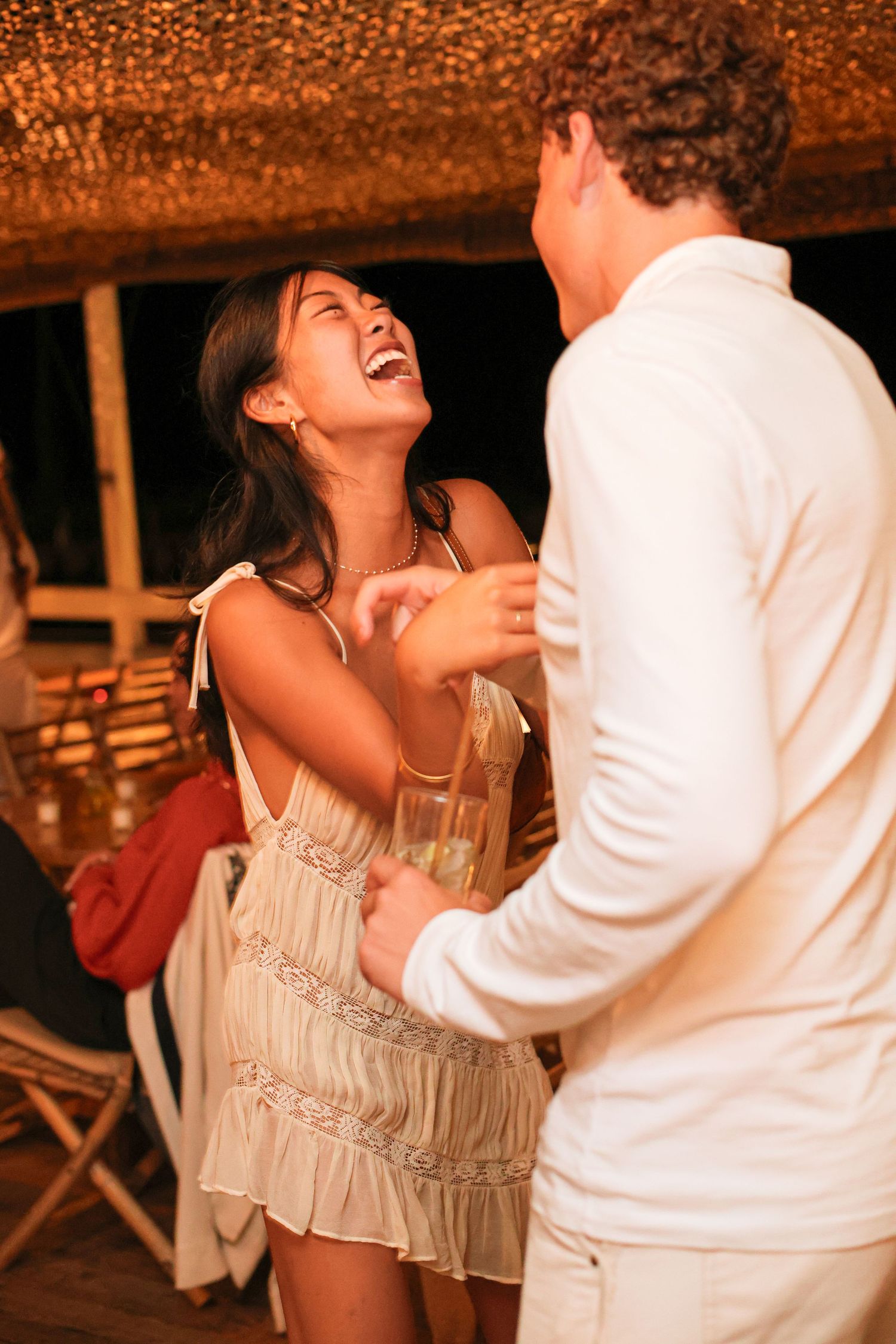 A laughing woman in a cream sundress dances with a man in a white shirt at a lively evening party under a thatched roof.