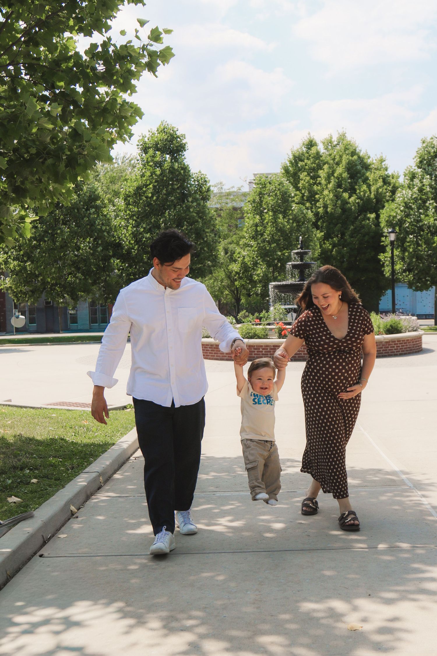 A young family with a toddler walks along a sunny park path, parents holding child's hands outdoors.