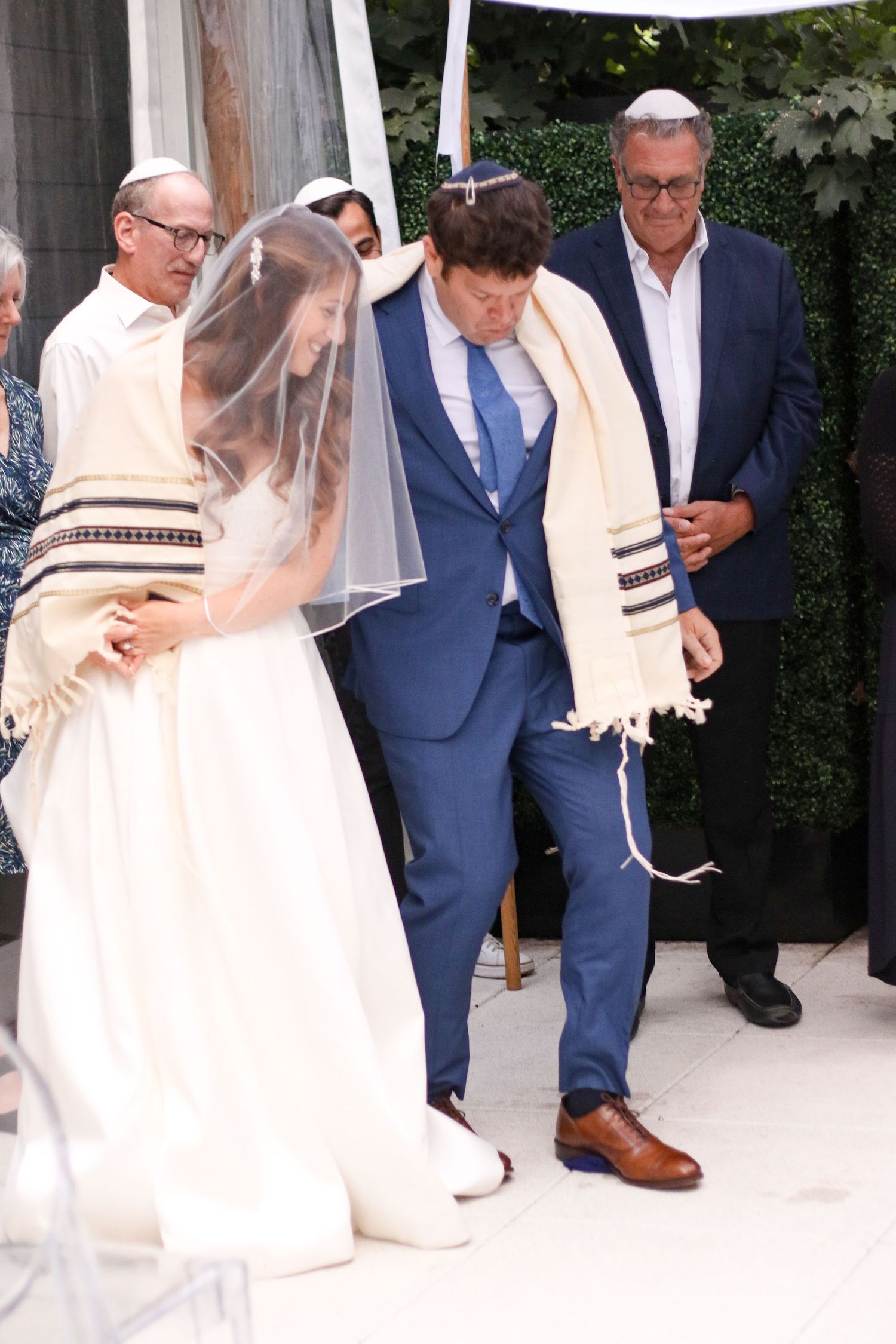 Jewish groom stomps the glass during traditional wedding ceremony under a chuppah with family.
