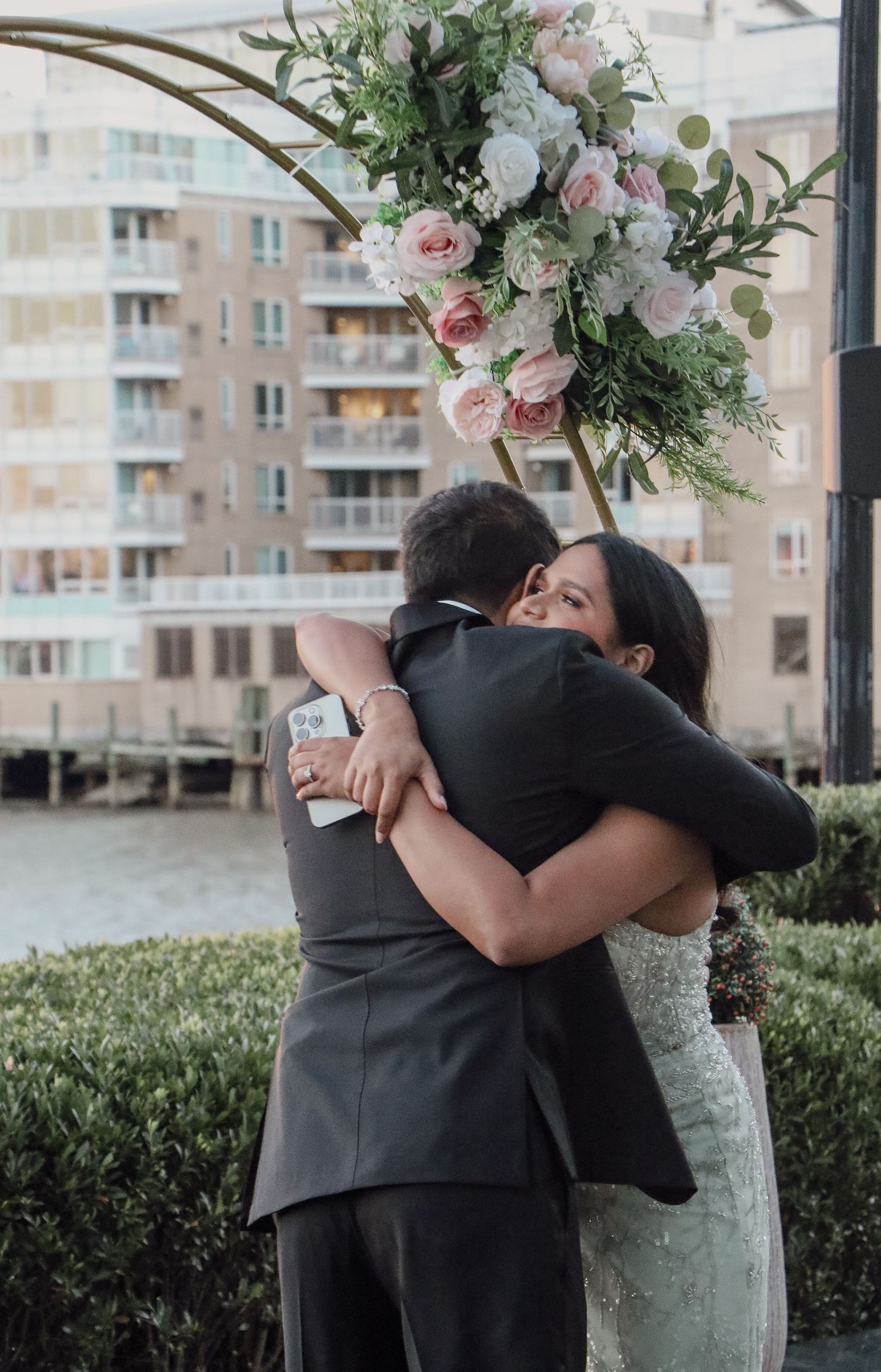 Newlyweds embrace in a heartfelt hug under a circular floral arch at a waterfront wedding.