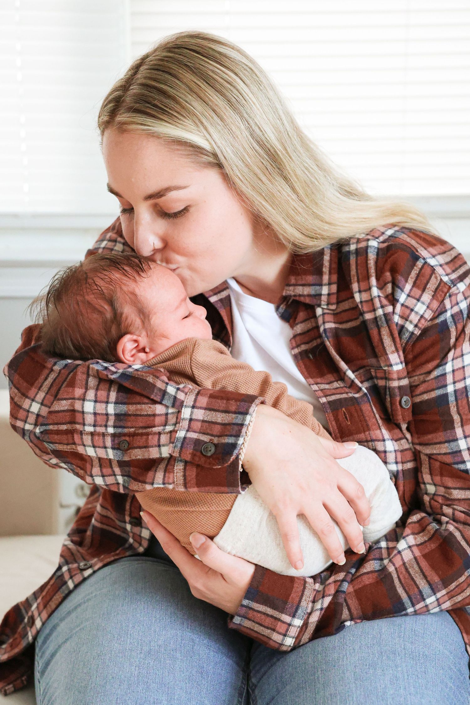 A blonde woman in a red plaid flannel shirt tenderly kisses the forehead of a sleeping newborn baby while cradling the infant in her arms.