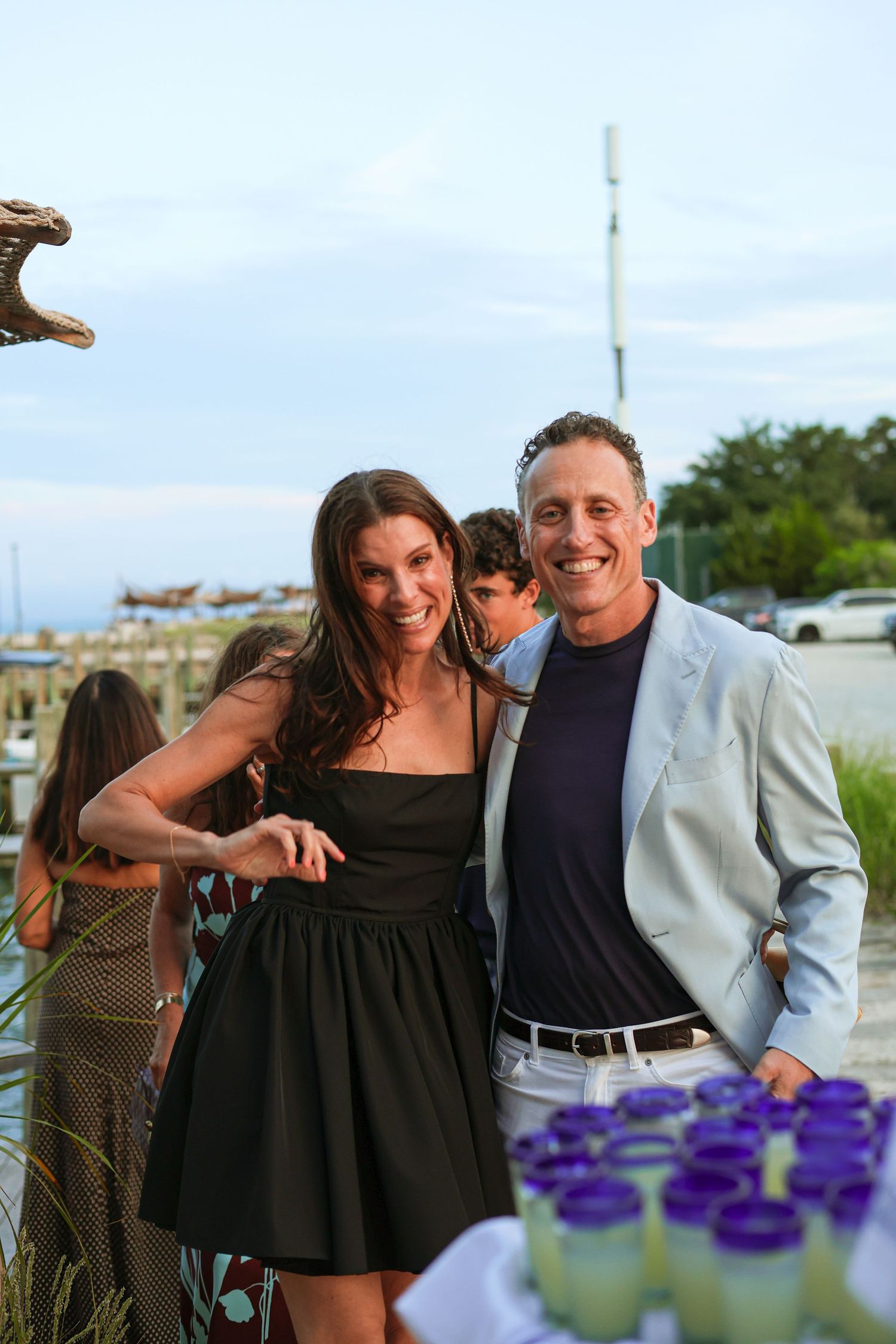Smiling woman in a black dress and man in a light blazer pose at an outdoor waterfront party with cups of lemonade in the foreground.