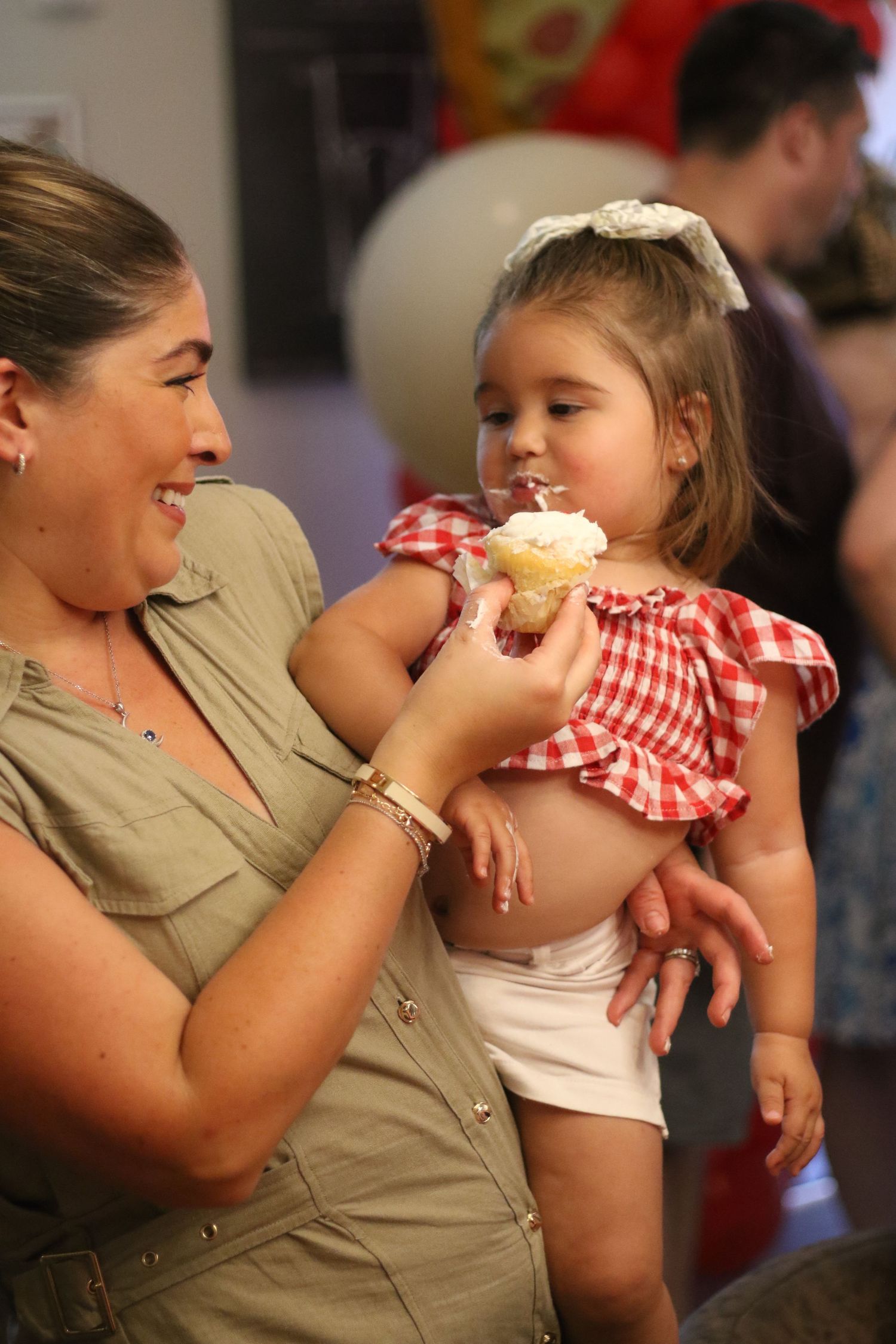 A smiling woman holds a toddler girl in a red gingham dress eating a cupcake at a birthday party.
