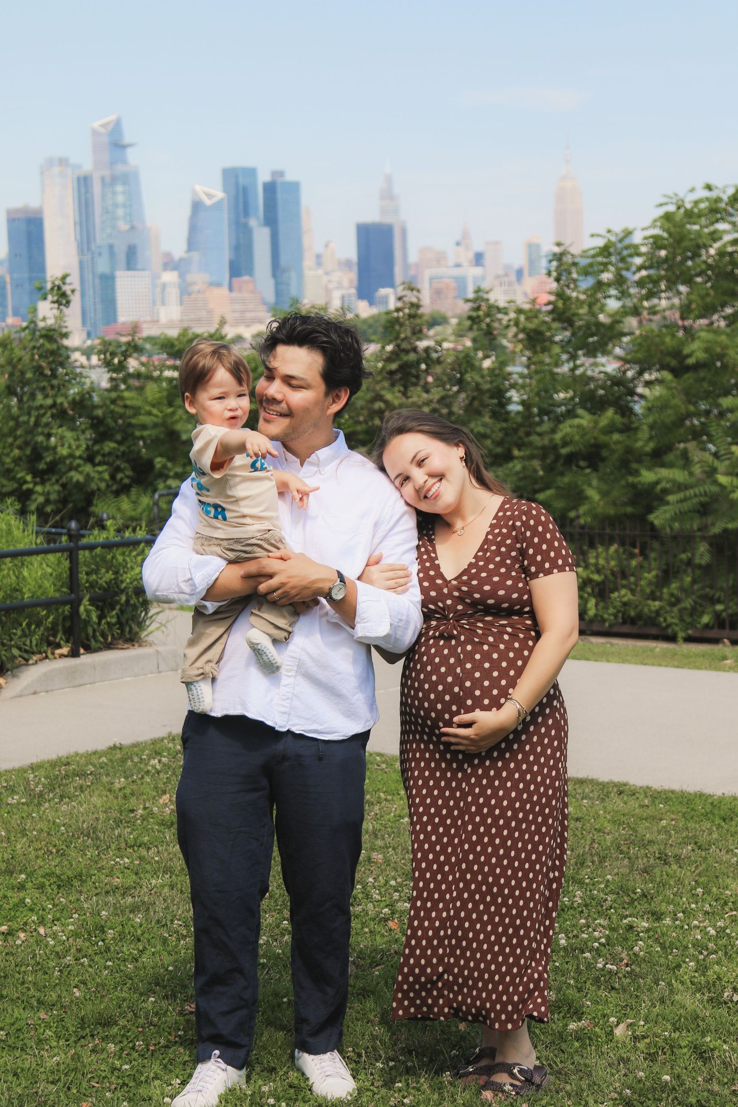 Happy family of three posing outdoors with NYC skyline backdrop, pregnant mother in brown polka dot dress.