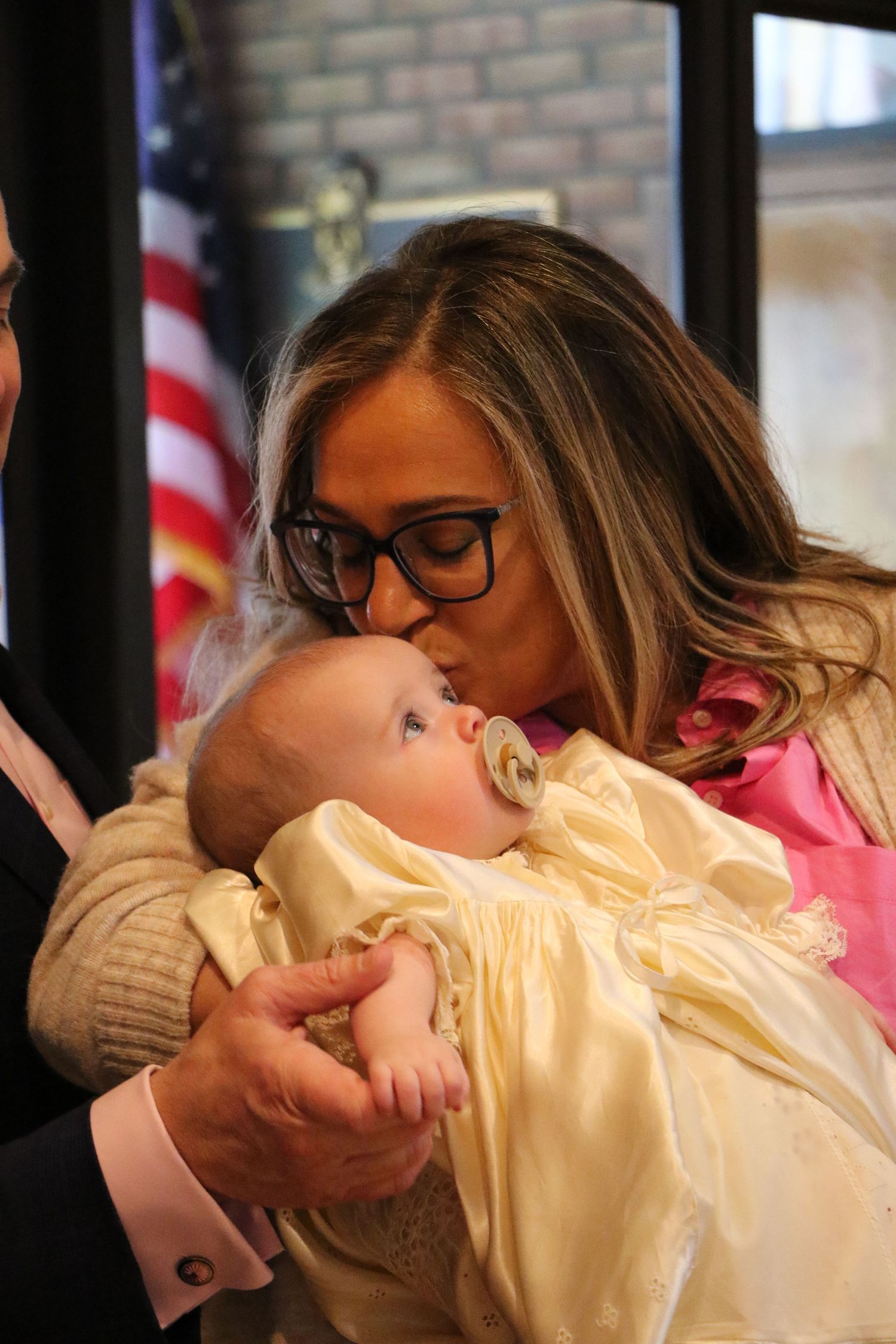 Woman with glasses kissing baby in christening gown near American flag.