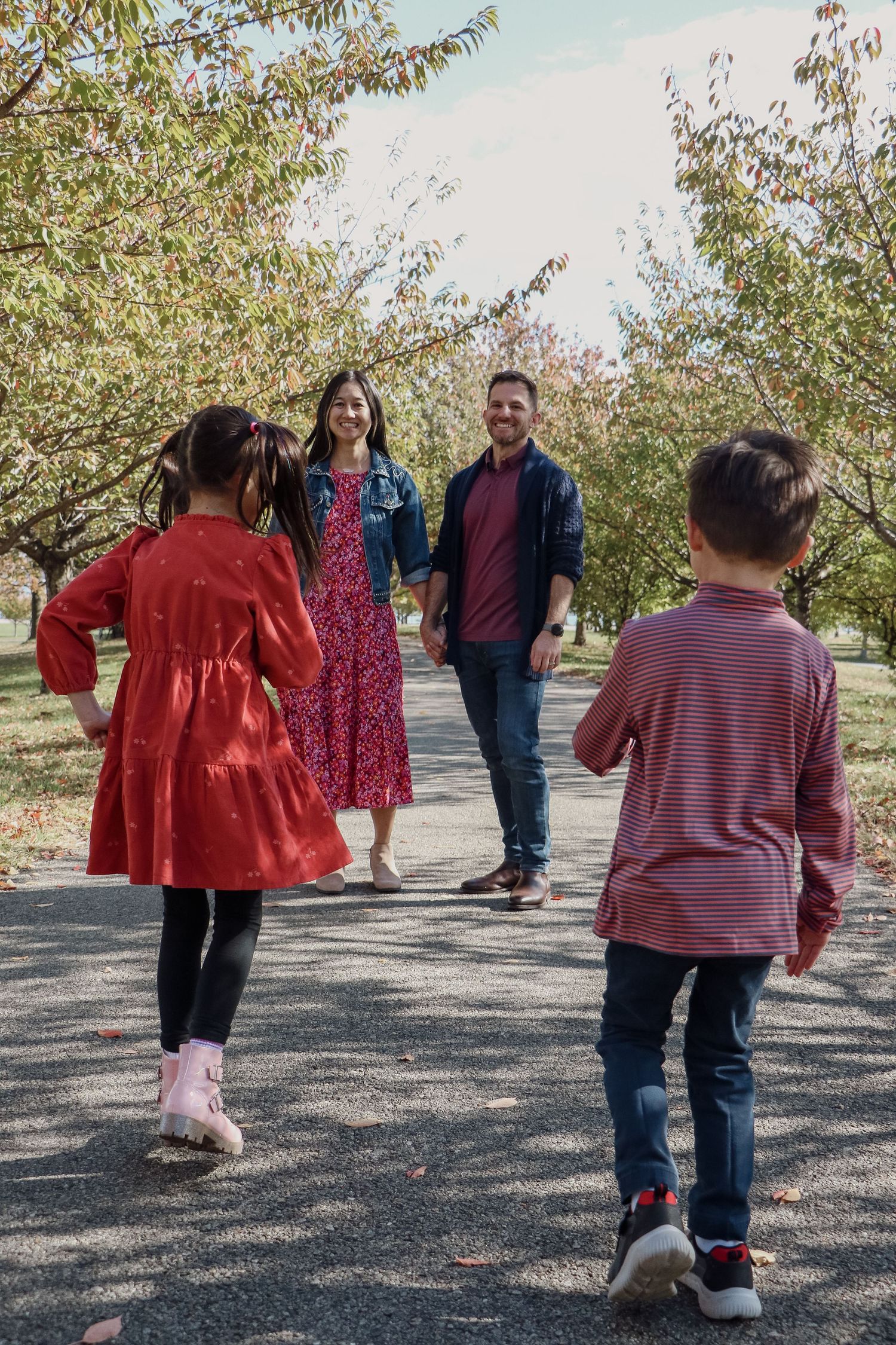 Family of four walks along tree-lined path in autumn, parents holding hands with two young children.