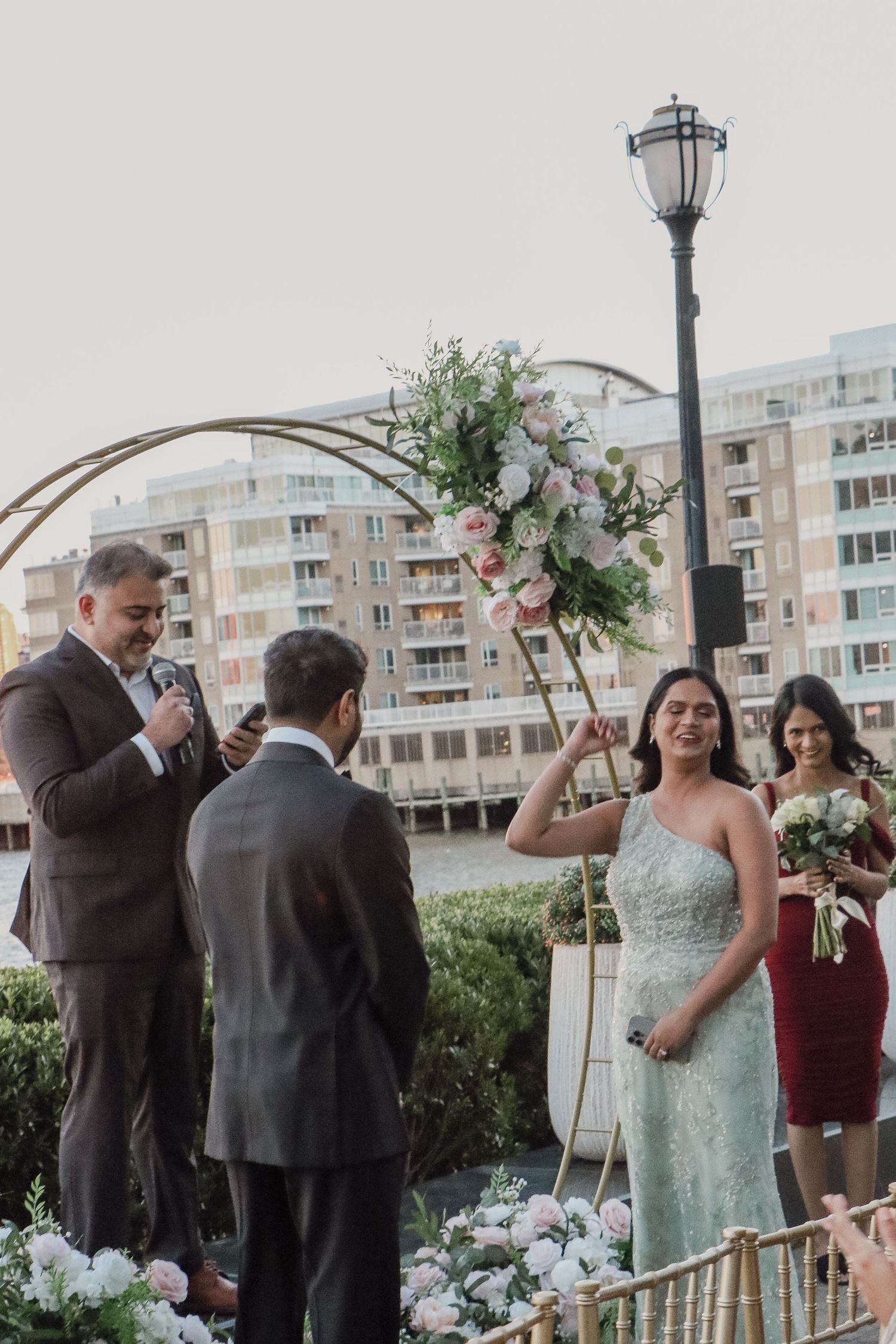 Bride in mint lace gown laughs joyfully during outdoor waterfront wedding ceremony at sunset.