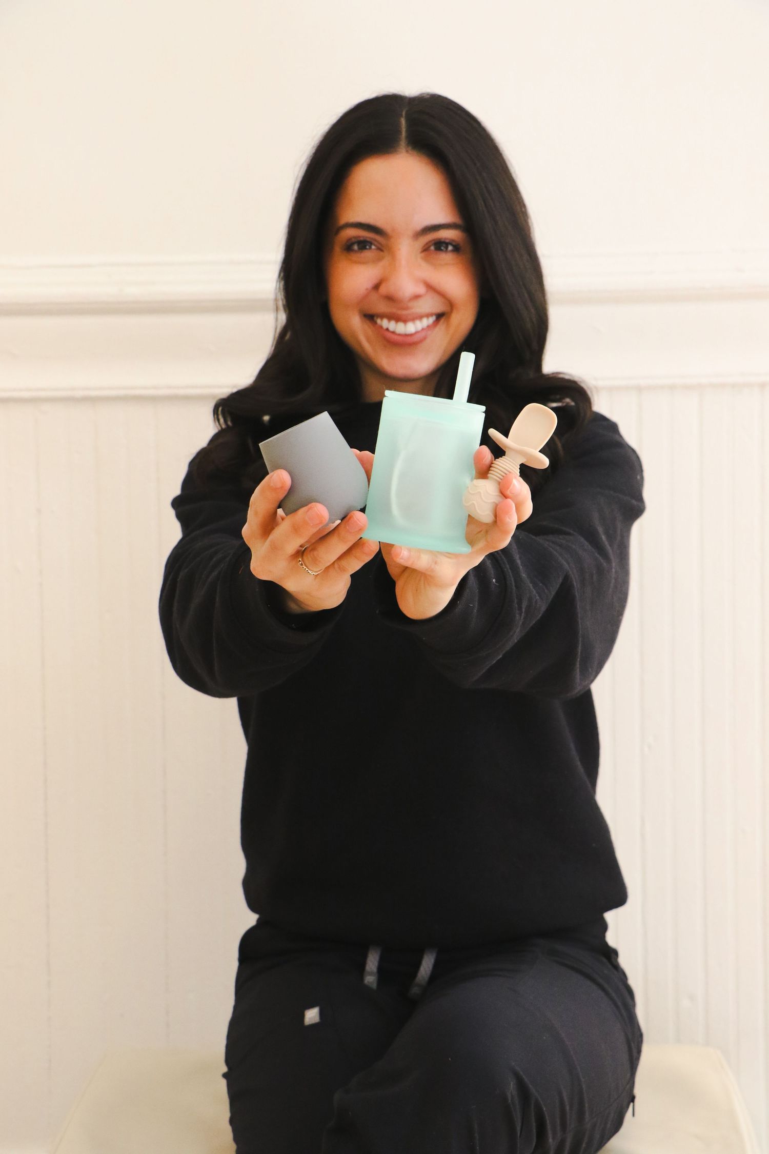 Smiling woman in black outfit holding mint green baby products and a wooden toy toward the camera.