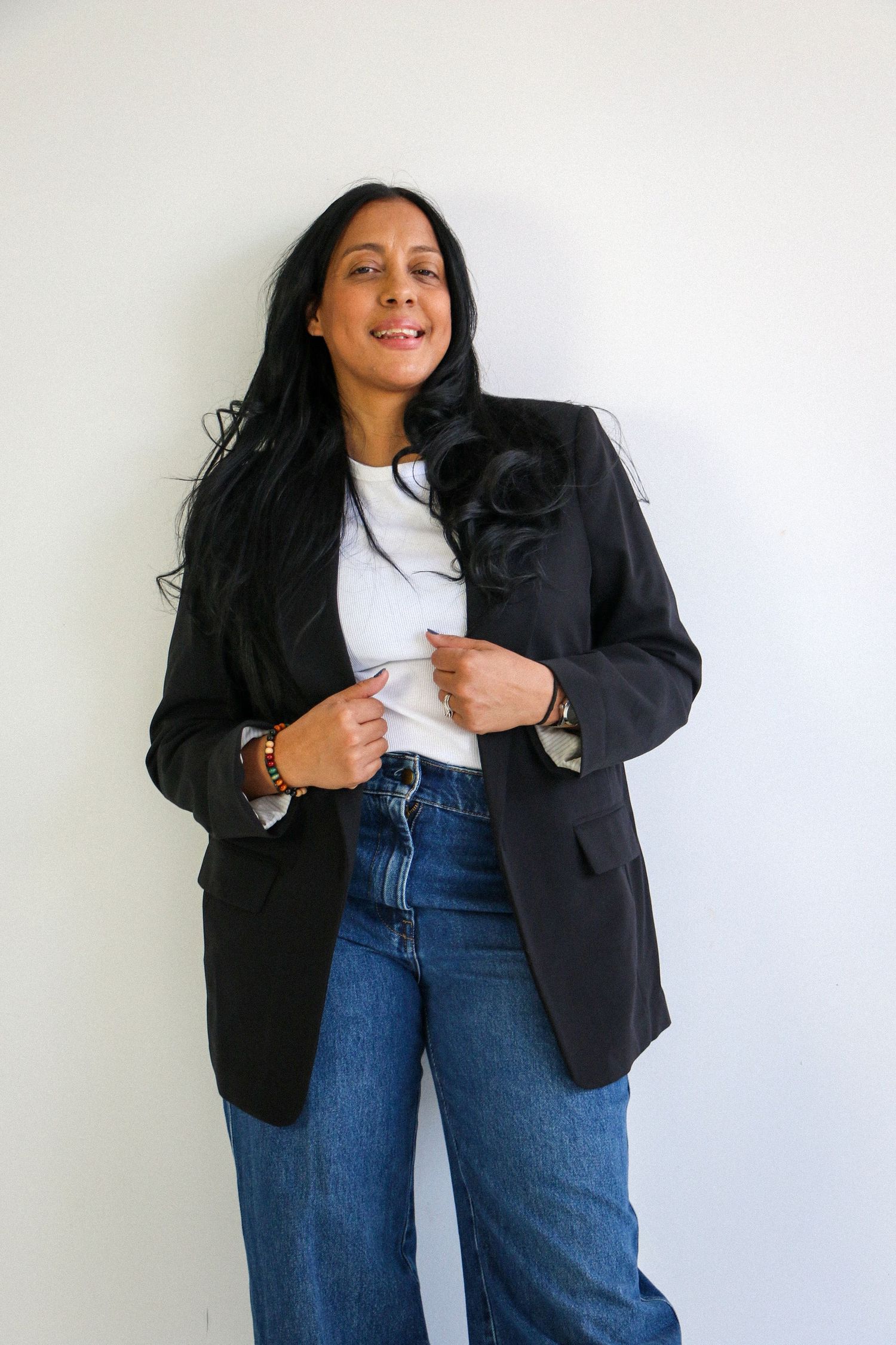 Woman in black blazer, white tee, and jeans posing confidently against a white wall.