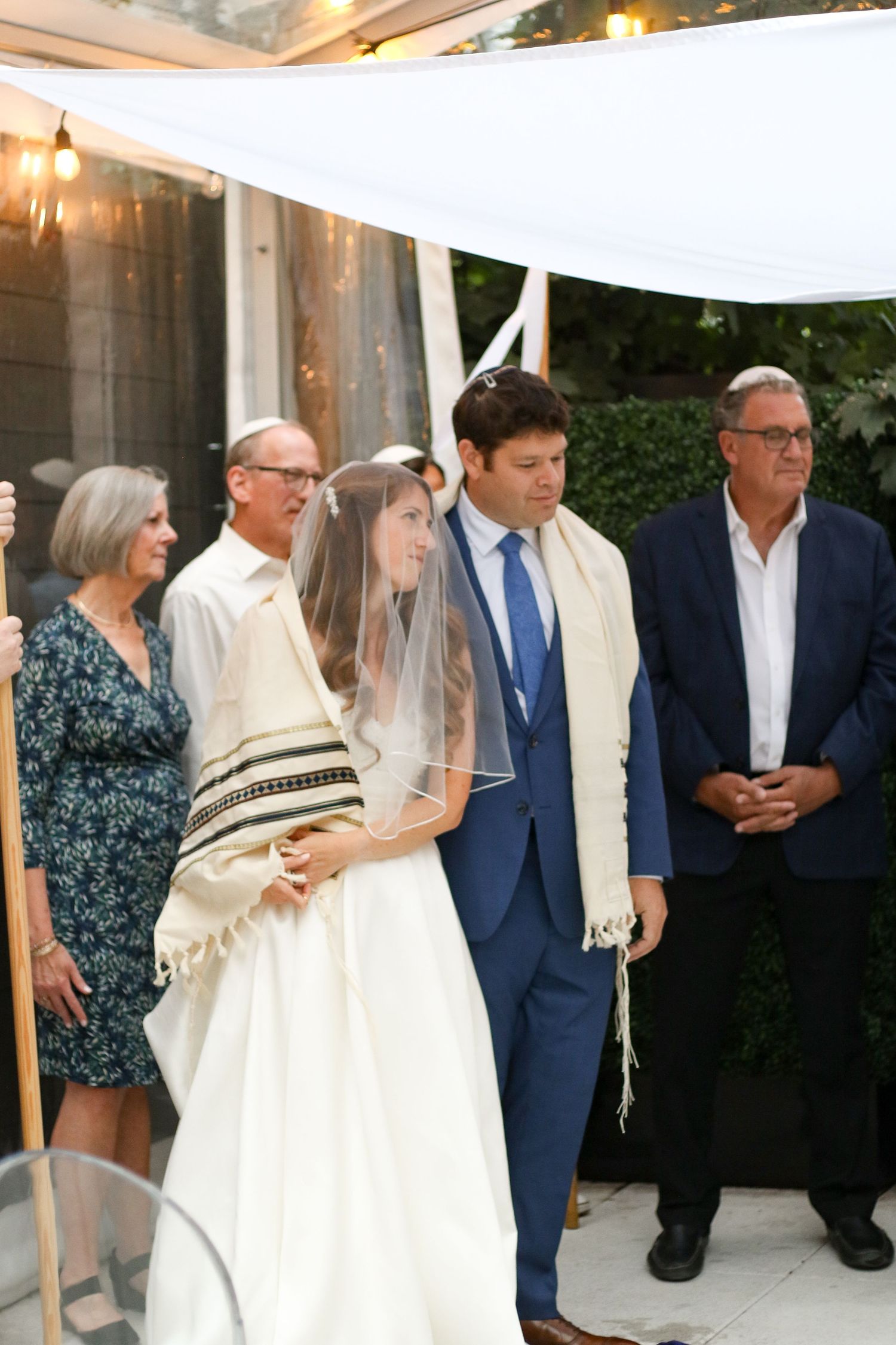 Jewish bride and groom stand under a white chuppah surrounded by family during wedding ceremony.