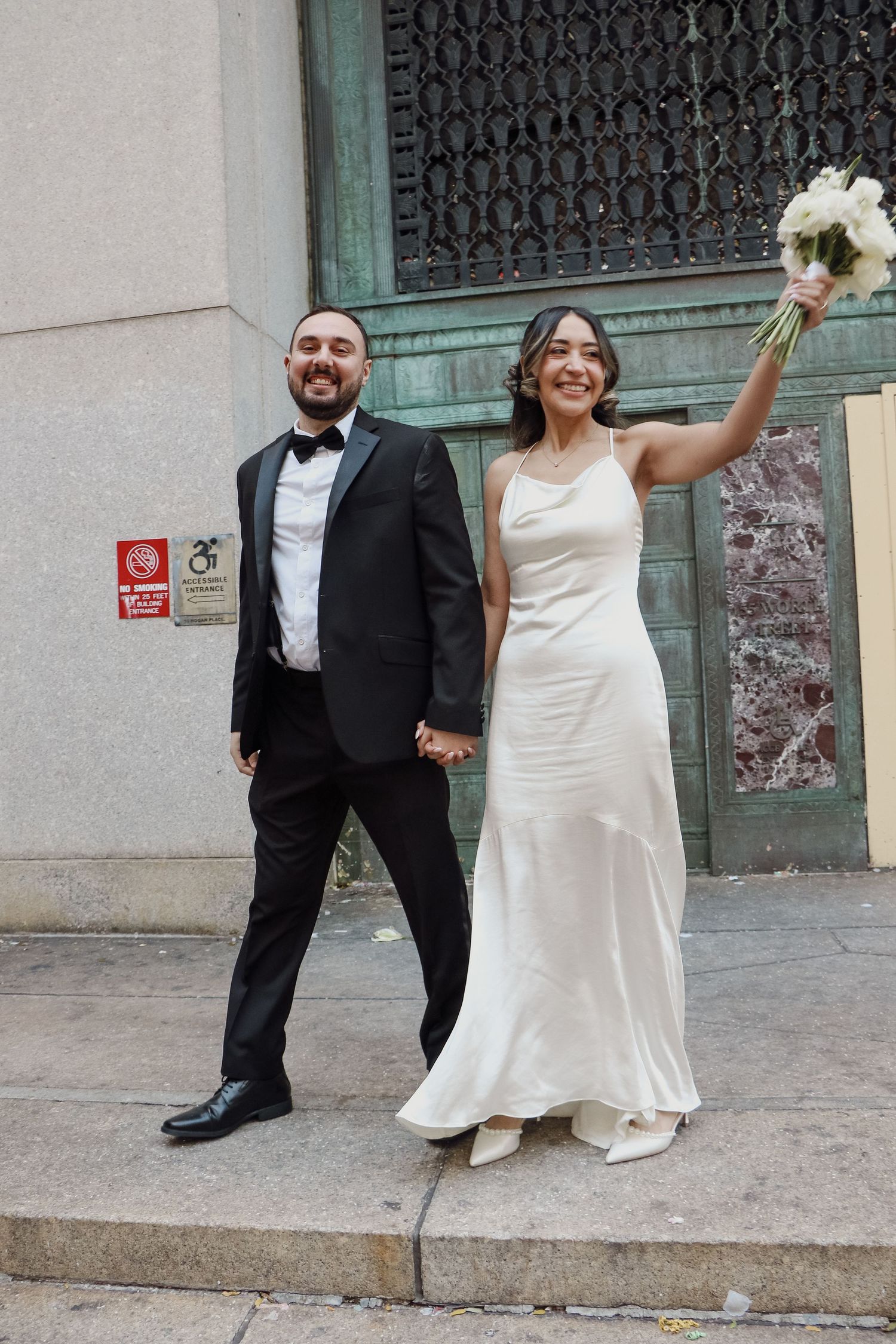 Joyful newlyweds walk outside NYC City Hall, bride in white satin gown raises bouquet, groom in black tuxedo.