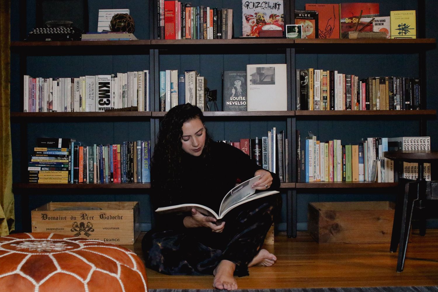 Young woman sitting cross-legged on floor reading a large book in front of a dark bookshelf.