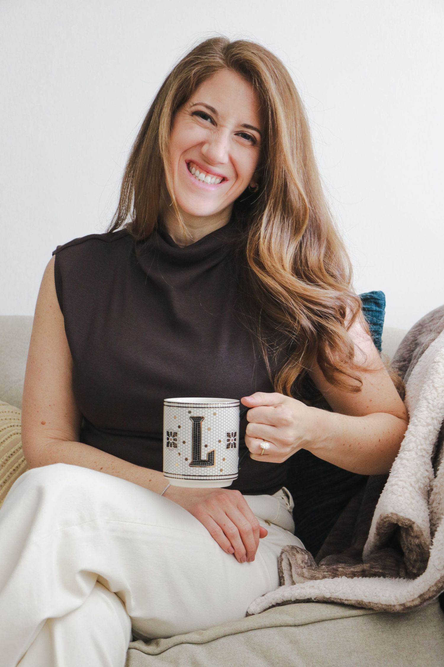 Cheerful woman holding monogram mug sitting on couch with cozy blanket.
