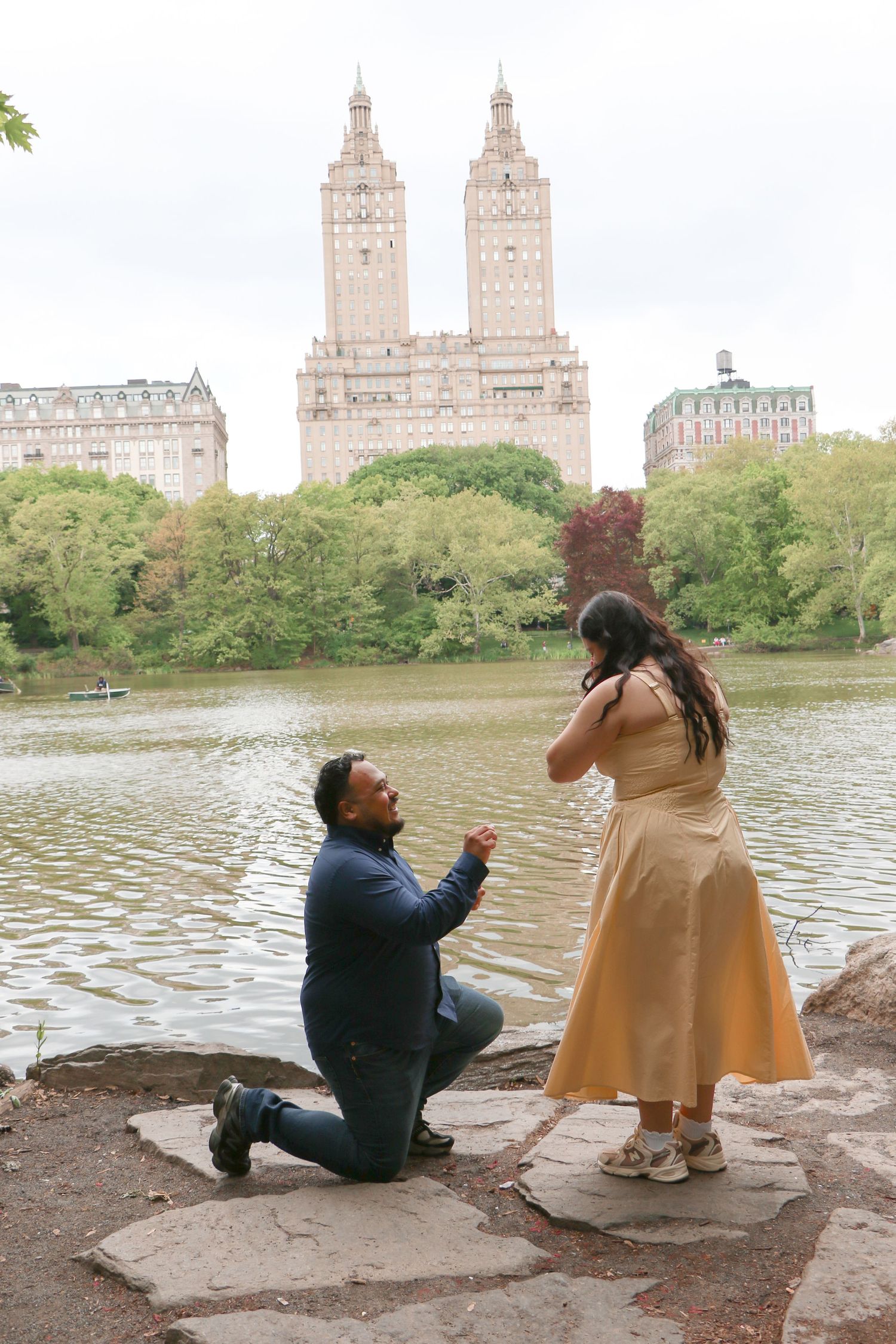 Man proposes on one knee to woman in gold dress by Central Park lake with iconic San Remo building backdrop.