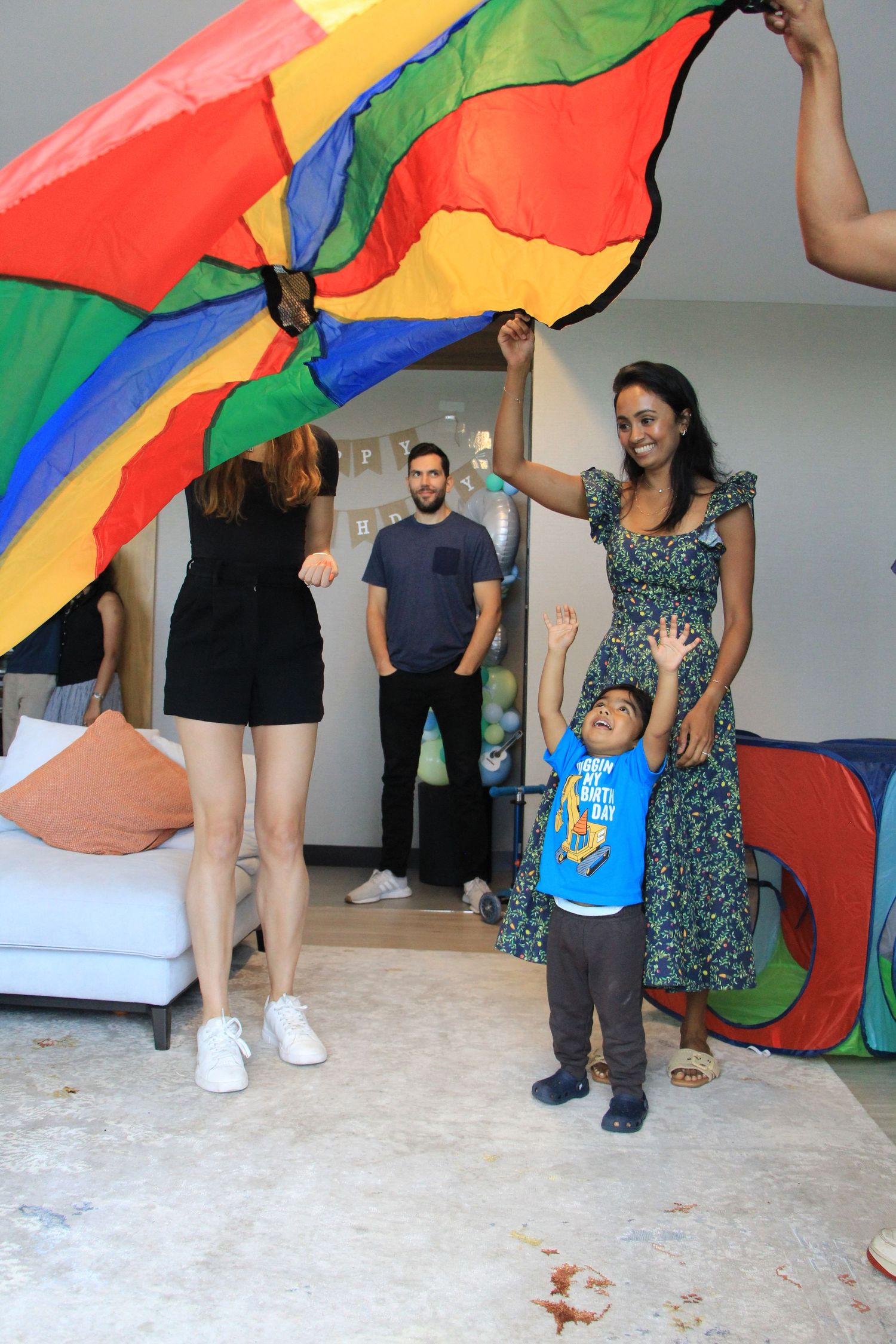 Family plays with a rainbow parachute with a birthday boy in a blue shirt at an indoor party.