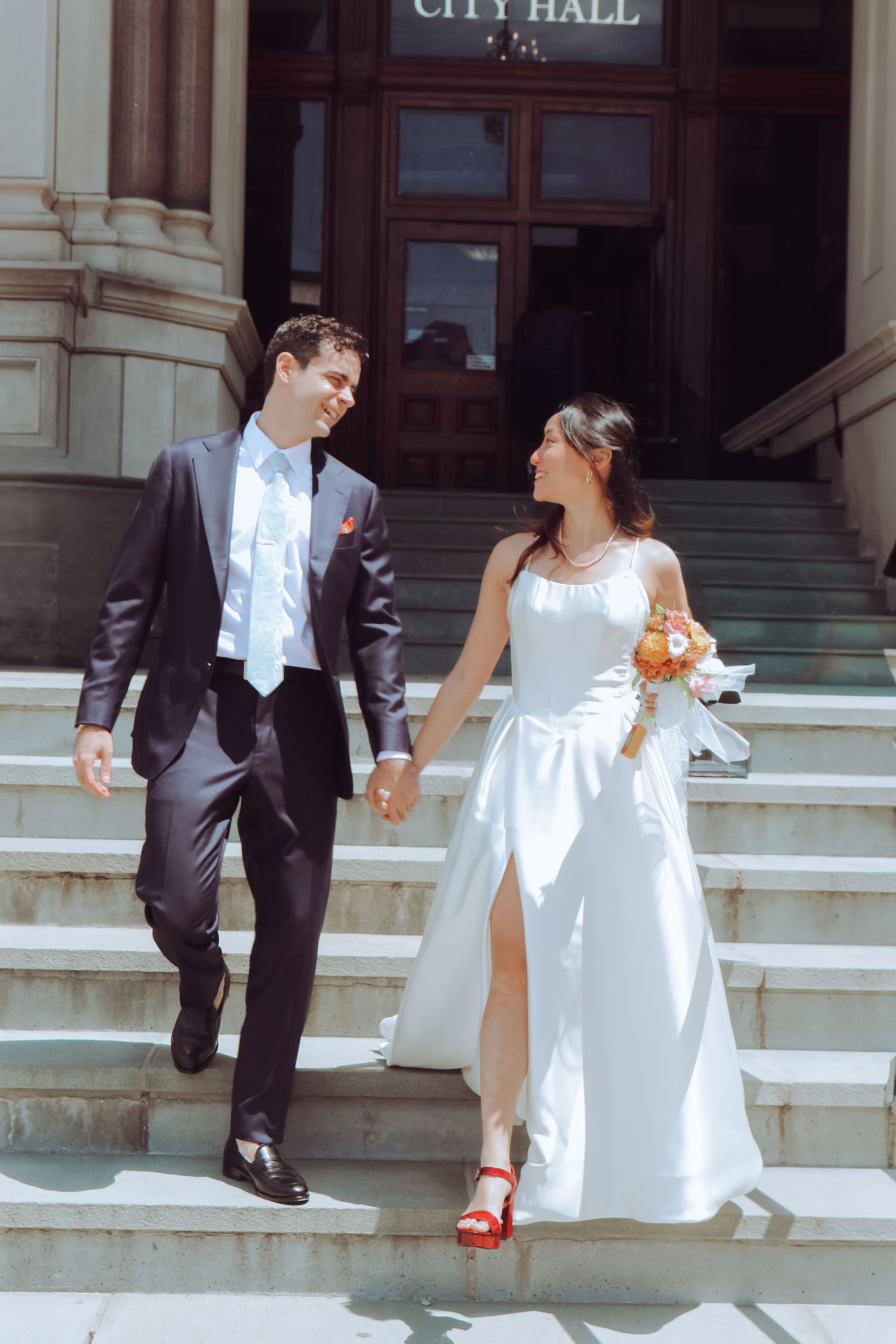 Newlywed couple holding hands walking down City Hall steps, bride in white gown with red heels, groom in dark suit.