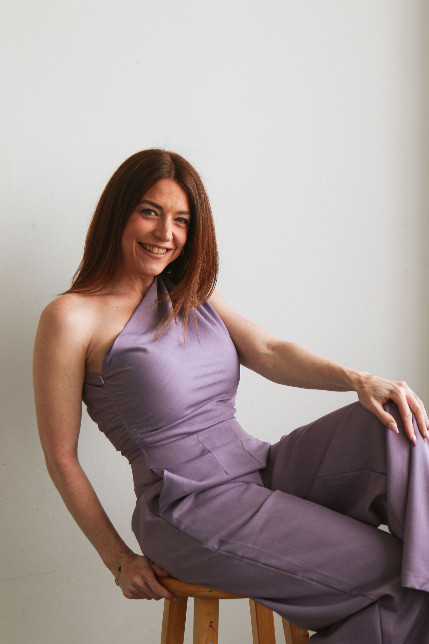 Woman in purple halter jumpsuit sitting on wooden stool, smiling confidently against light gray background.